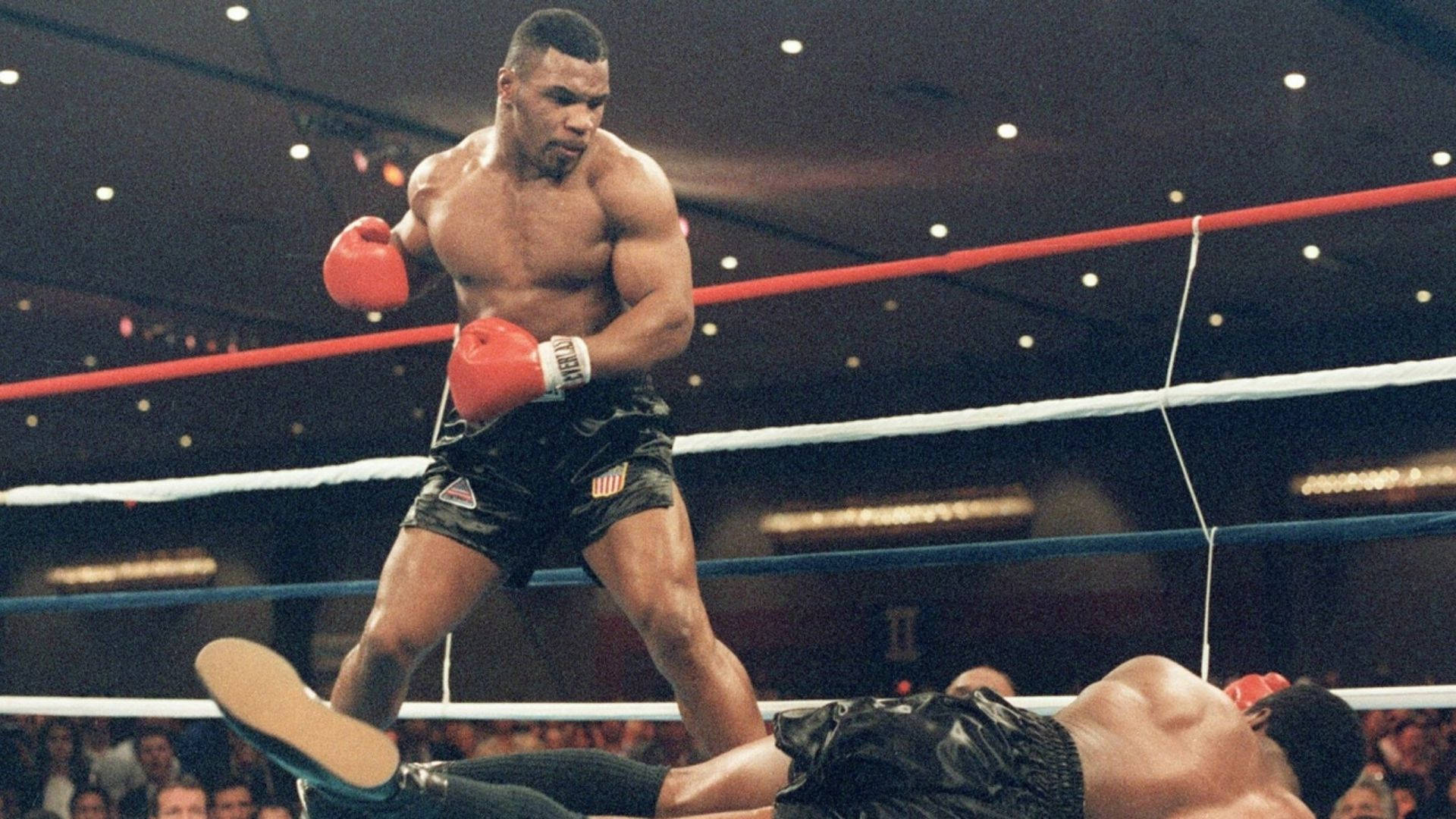 麥克·泰森 桌布 壁紙 — Mike Tyson 在擂台上勝利地站著，而他的對手在明亮的場館燈光下被打敗躺在地上。