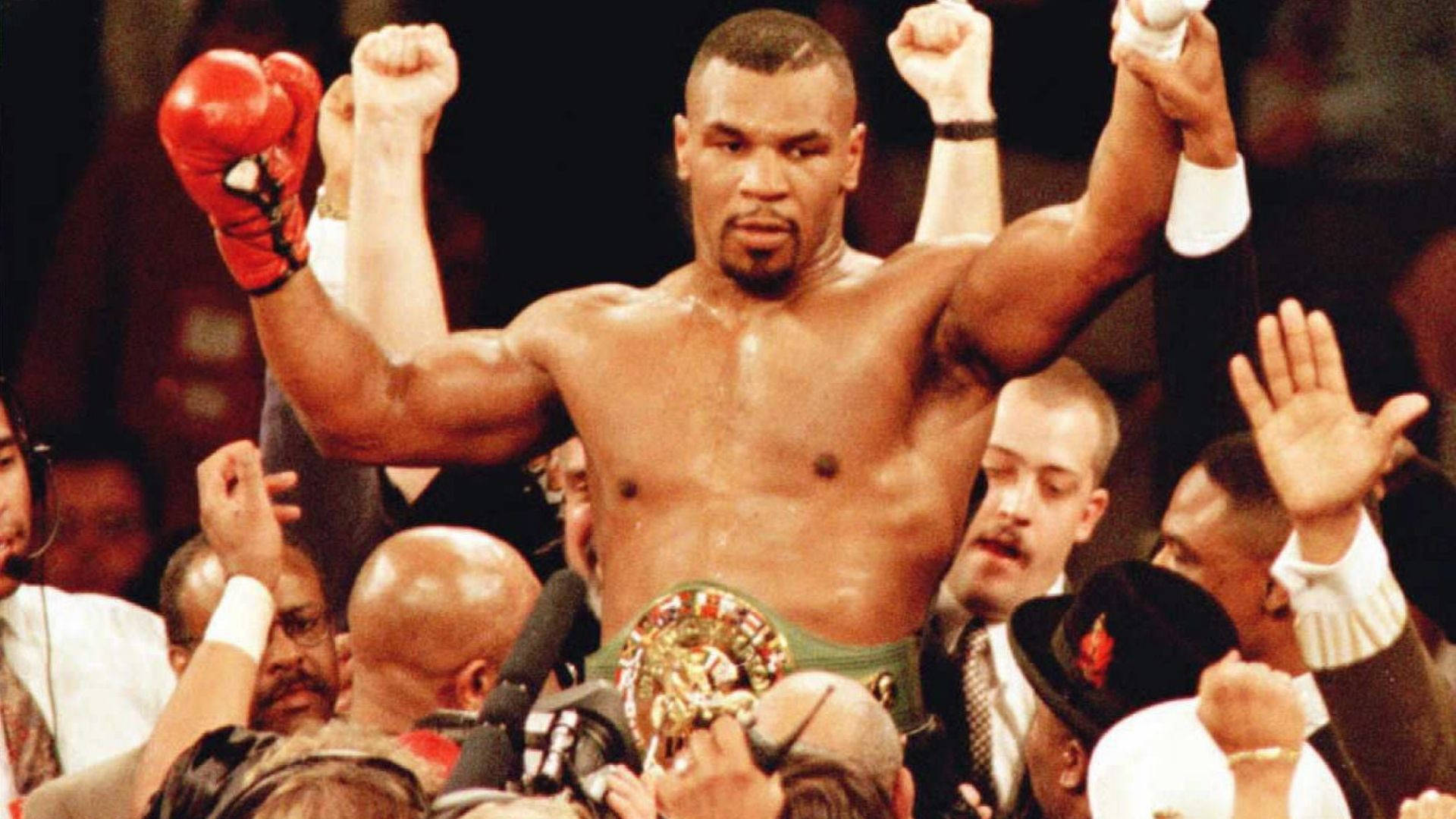 麥克·泰森 桌布 壁紙 — Mike Tyson 在擂台上勝利地高舉拳頭，戴著紅色手套和冠軍腰帶，被慶祝的人群包圍。