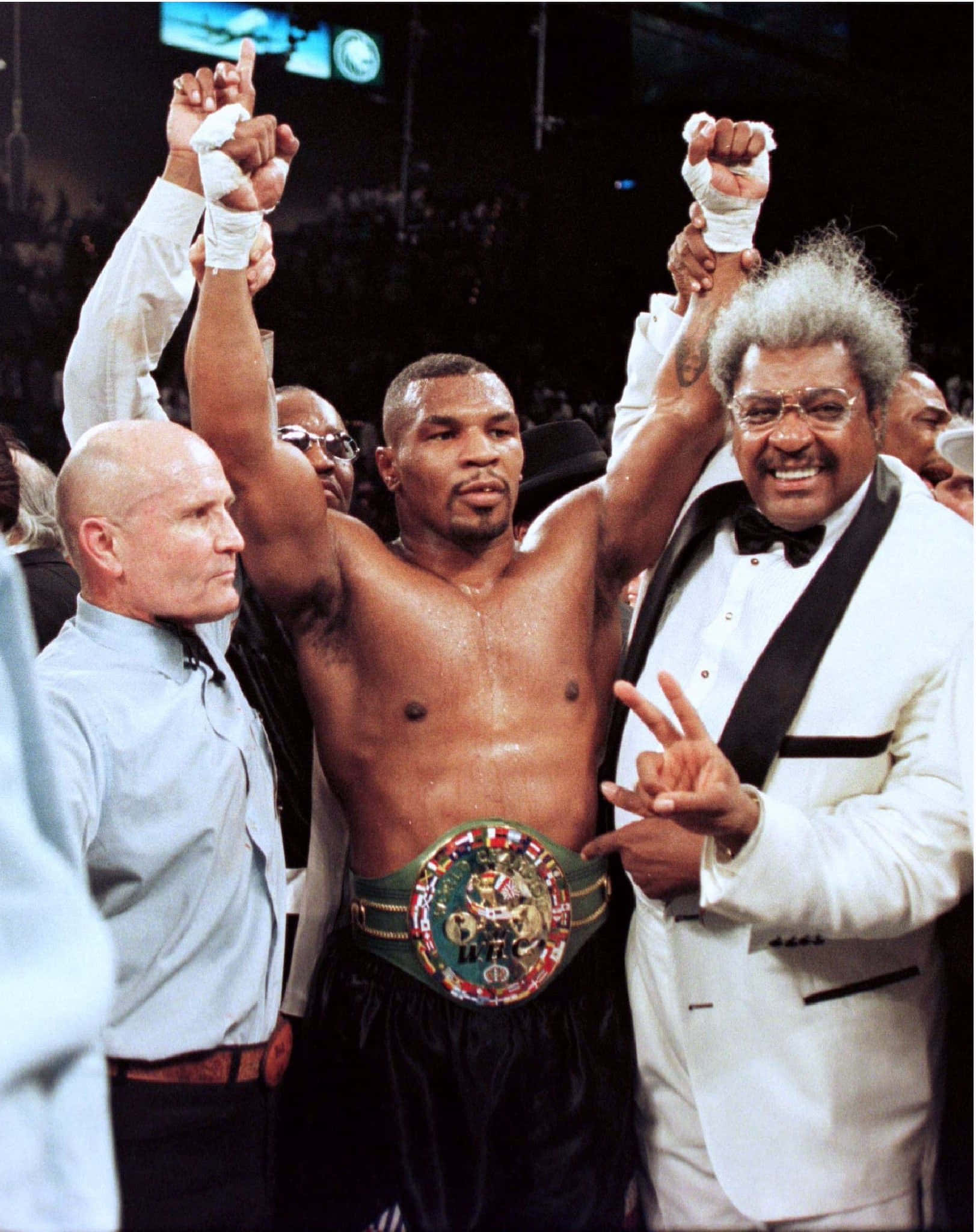麥克·泰森 桌布 壁紙 — Mike Tyson 在擂台上高舉拳頭和冠軍腰帶慶祝勝利，官員和一位身穿燕尾服微笑的男子站在他兩旁。