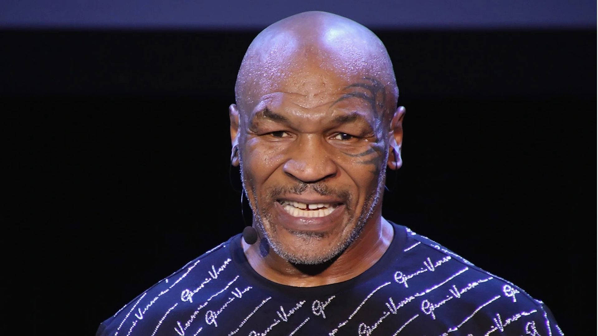 麥克·泰森 桌布 壁紙 — Mike Tyson 在舞台上燦爛地笑著，穿著 Gucci 襯衫，在聚光燈下，背景為黑色。