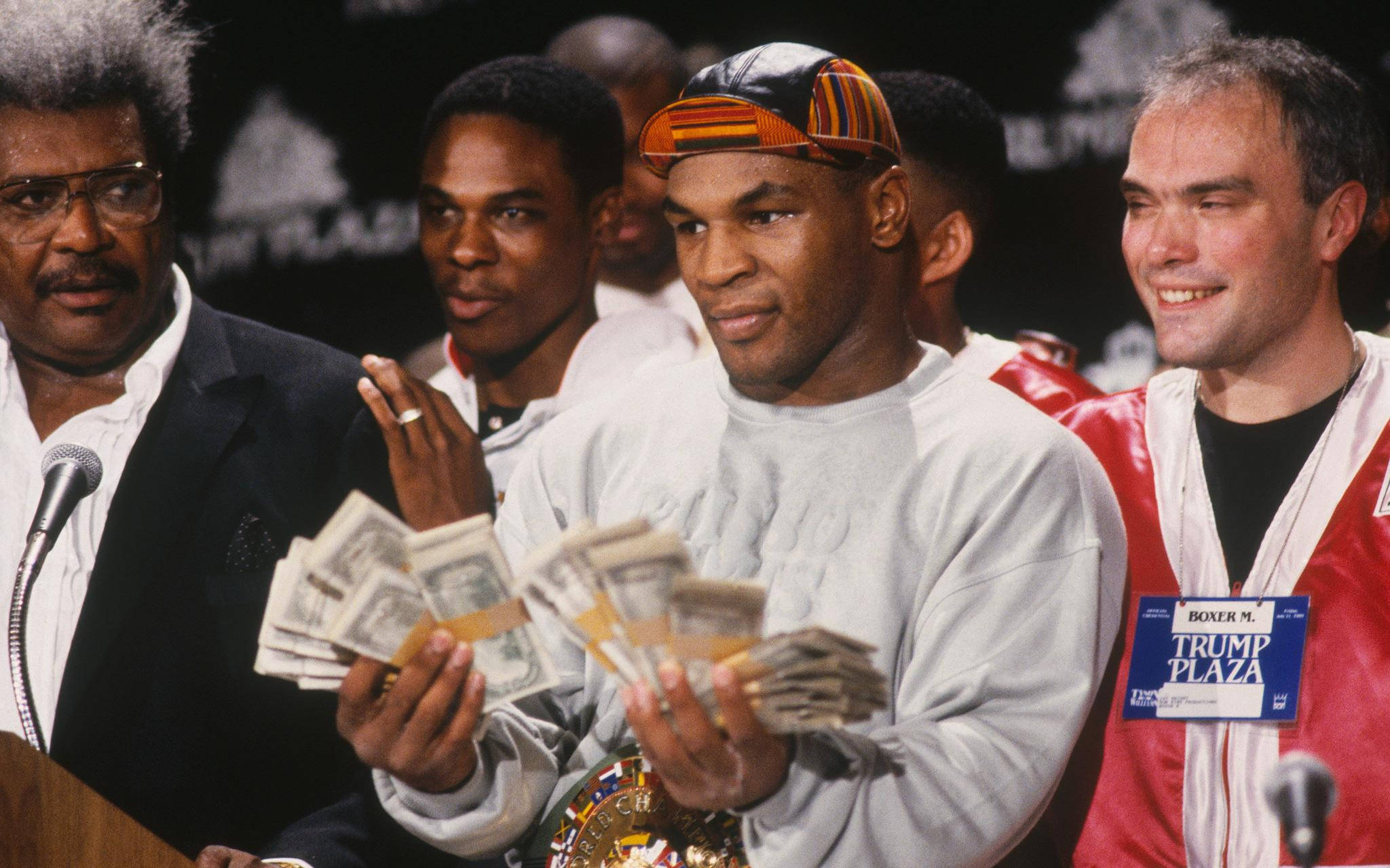 麥克·泰森 桌布 壁紙 — Mike Tyson 在記者會上手持一疊疊現金和冠軍腰帶，周圍有微笑的官員。