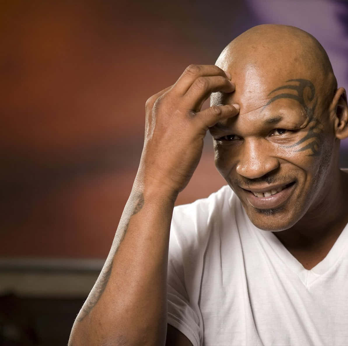 麥克·泰森 桌布 壁紙 — Mike Tyson 溫暖地微笑，同時用刺青的手觸摸額頭，背景是模糊的暖色調。