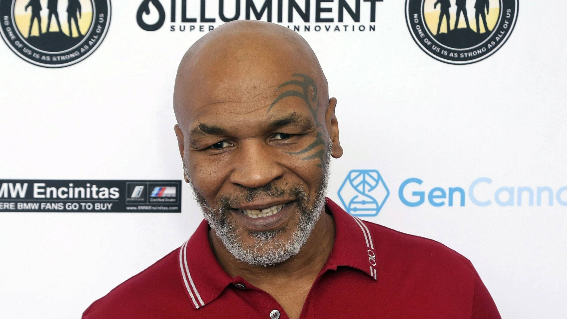 麥克·泰森 桌布 壁紙 — Mike Tyson 穿著紅色Polo衫，在品牌活動背景前溫暖地微笑。