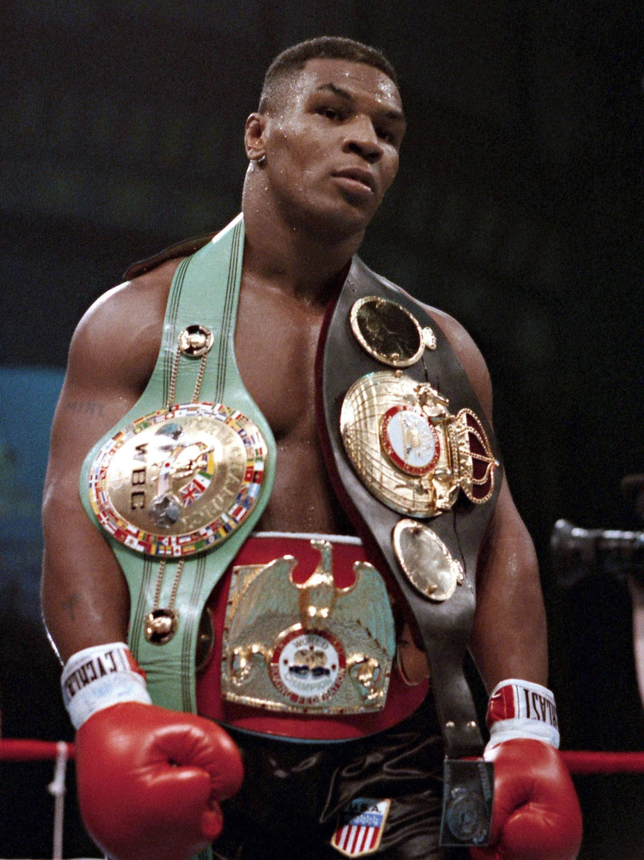 麥克·泰森 桌布 壁紙 — Mike Tyson 站在拳擊擂台上，佩戴多條冠軍腰帶和紅色手套，表情專注。