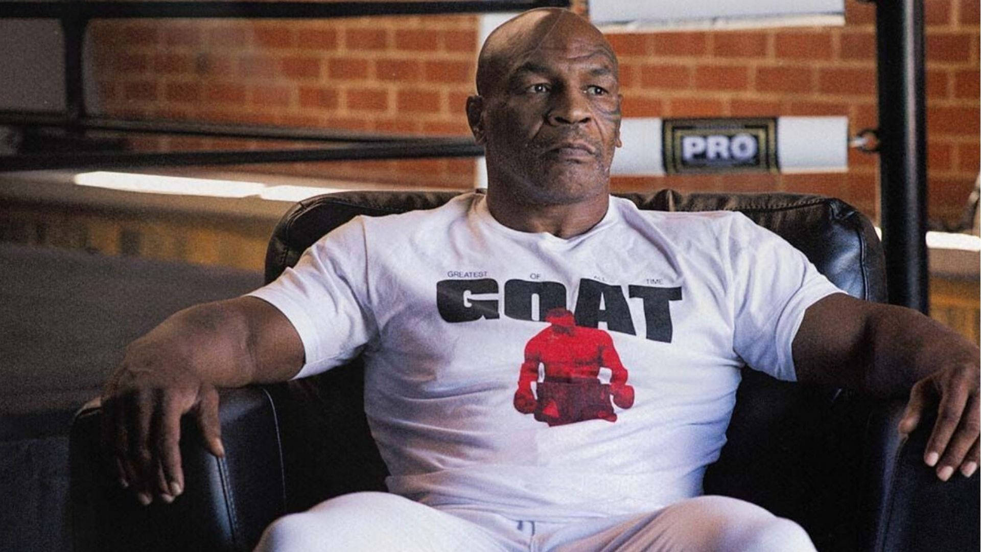 麥克·泰森 桌布 壁紙 — Mike Tyson 自信地坐在黑色皮椅上，穿著一件印有 'GOAT' 和紅色拳擊人物圖案的白色 T 恤。