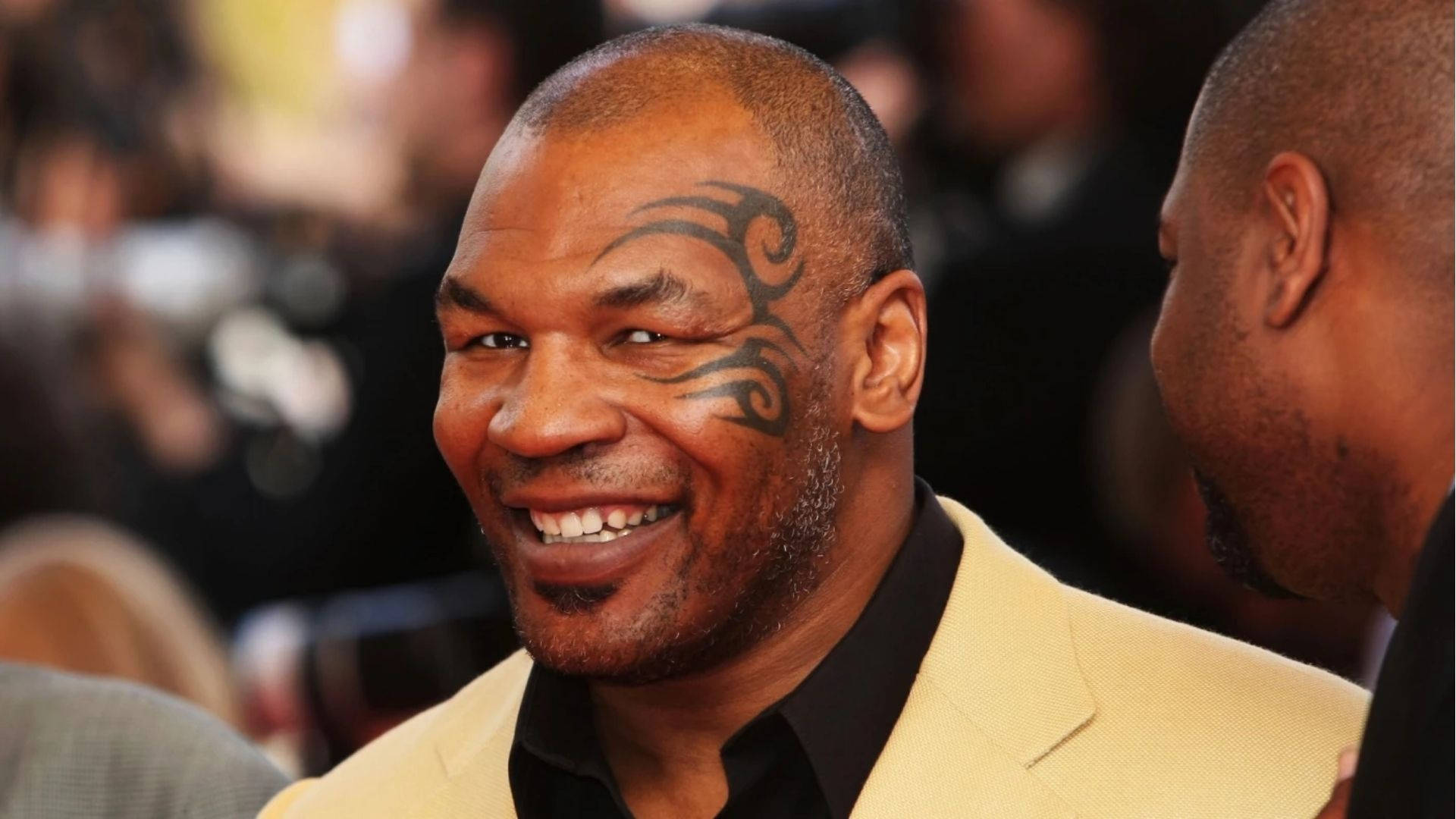 麥克·泰森 桌布 壁紙 — Mike Tyson 露出燦爛笑容，臉頰上可見其標誌性的部落紋身，身穿米色西裝。