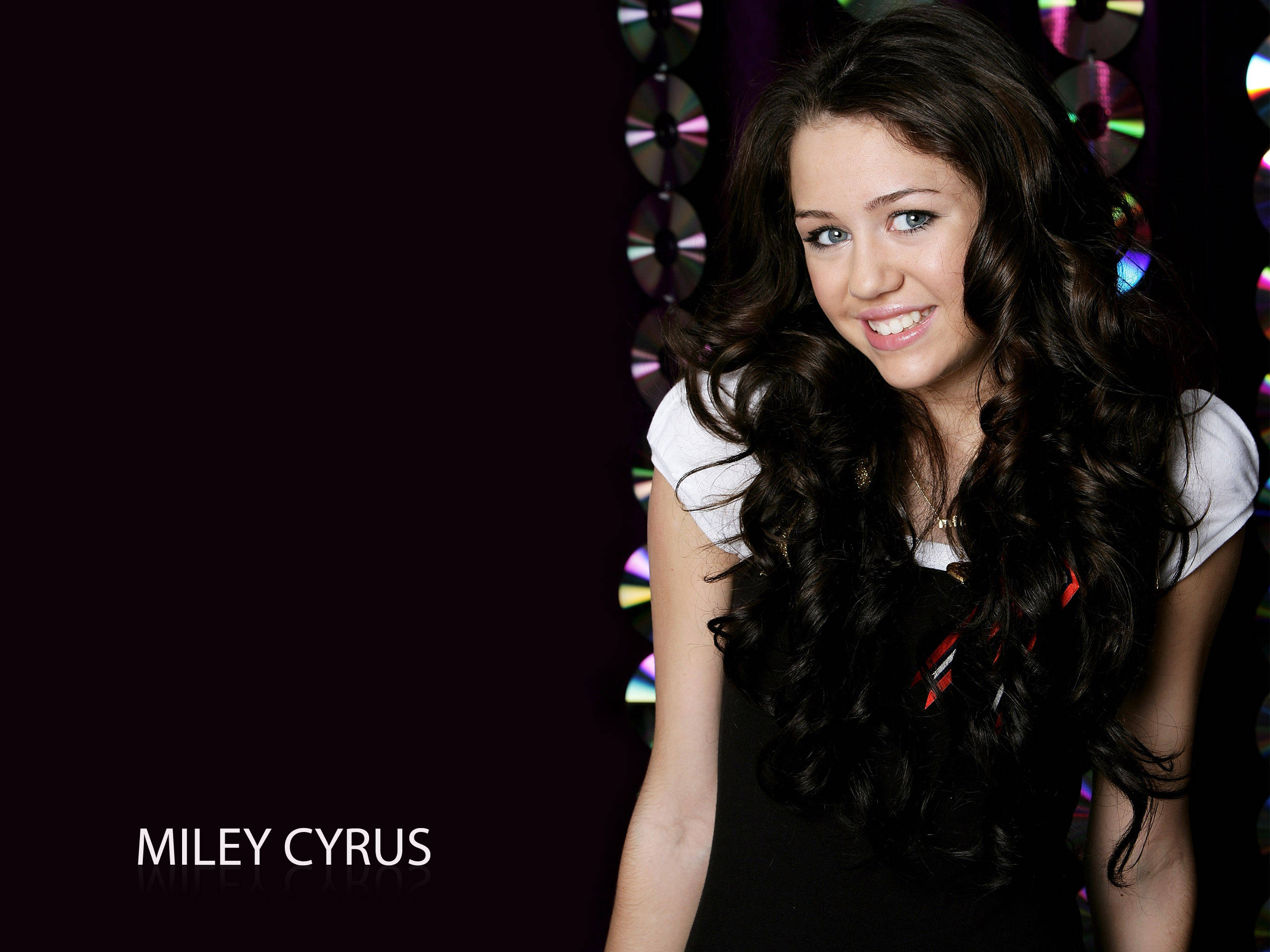 miley 桌布 壁紙 — Miley Cyrus 在黑暗背景下露出燦爛笑容，身後有色彩繽紛的CD反射。