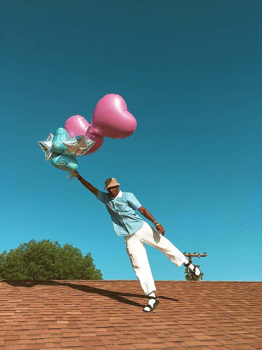 Tyler The Creator Aesthetic 桌布 壁紙 — 一名身穿淺藍色襯衫和白色褲子的男子在紅色瓦片屋頂上保持平衡，手持粉紅色心形和星形氣球，背景是清澈的藍天。