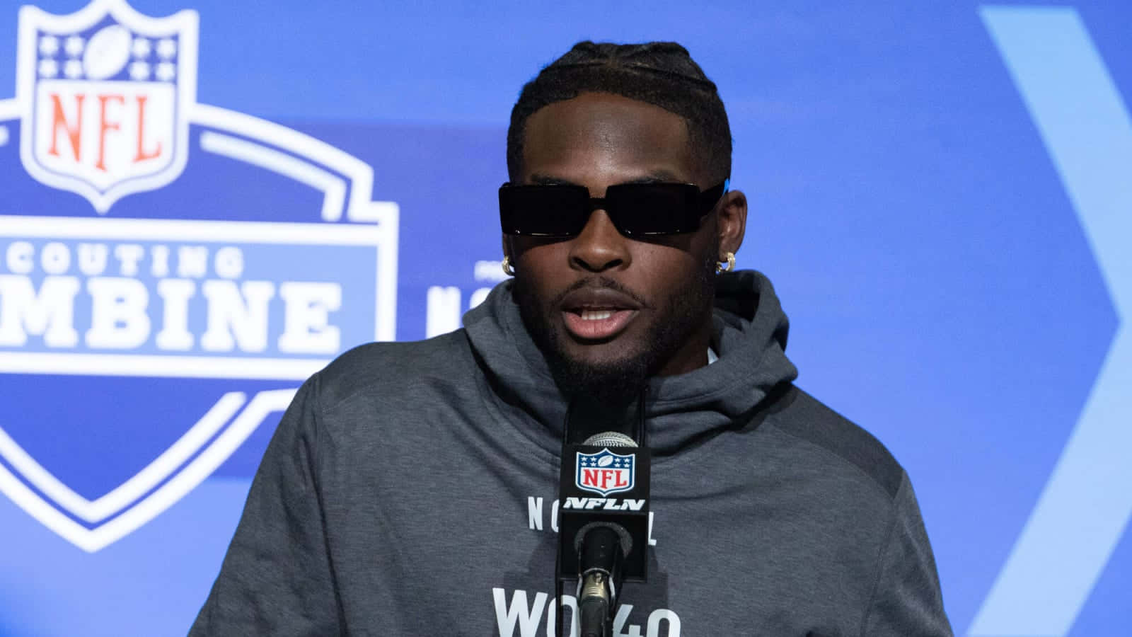  壁紙 — 一名戴著太陽眼鏡和灰色連帽衫的美式足球員在NFL選秀聯合試訓記者會上對著麥克風說話。
