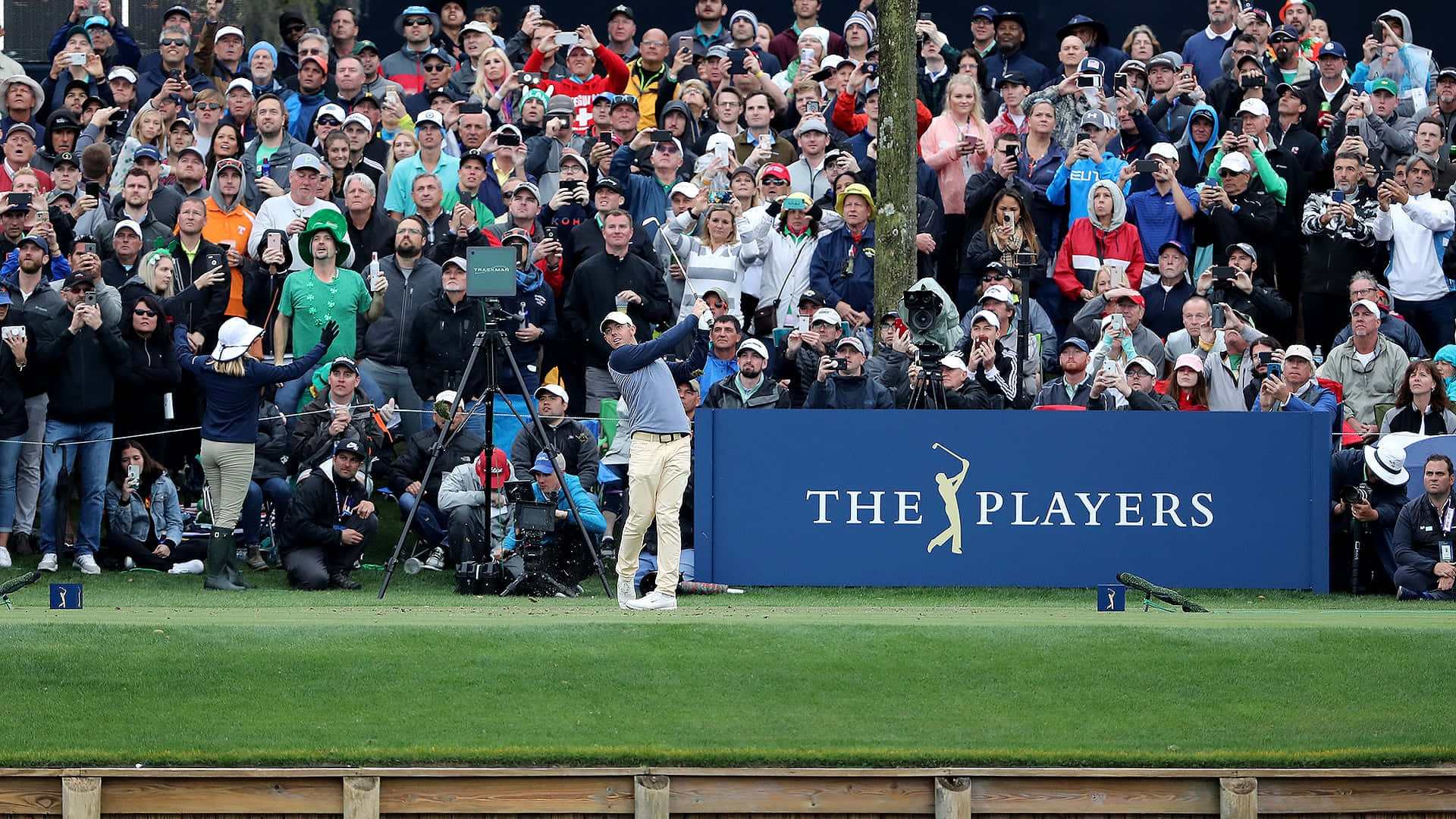 The Players Championship 桌布 壁紙 — 一名高爾夫球手在球員錦標賽的翠綠球道上揮桿，身後藍色品牌圍欄後擠滿歡呼的觀眾。