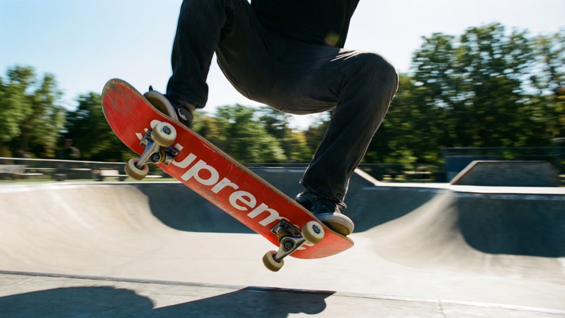 Supreme Skateboard Action 壁紙 — 一名滑板手在半空中，於陽光明媚的混凝土滑板公園坡道上，用紅色Supreme滑板表演技巧。