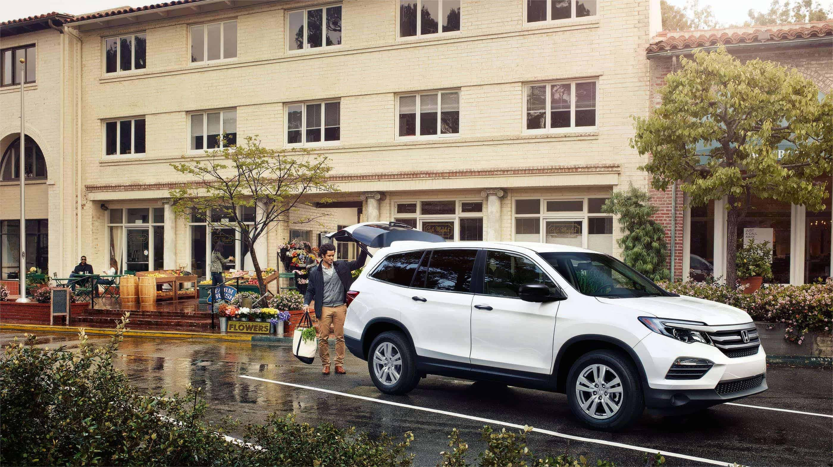 Honda Pilot 桌布 壁紙 — 一名男子站在一輛白色SUV旁邊，後車廂打開，位於潮濕的停車場，附近有花店和米色建築物。