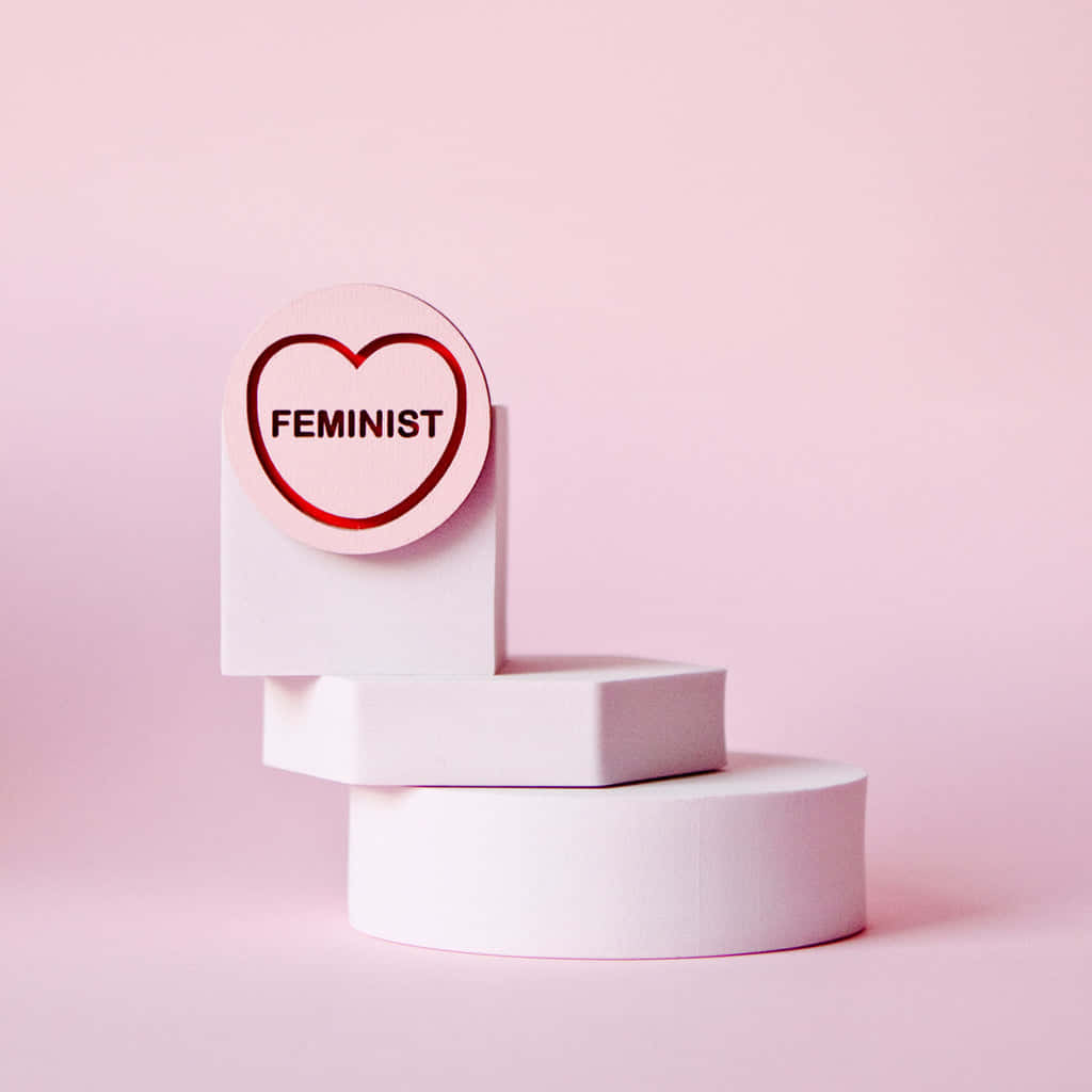  壁紙 — 一個極簡風格的粉紅色心形標誌，上面寫著「feminist」，放置在白色幾何形狀的積木上，背景是柔和的粉紅色。
