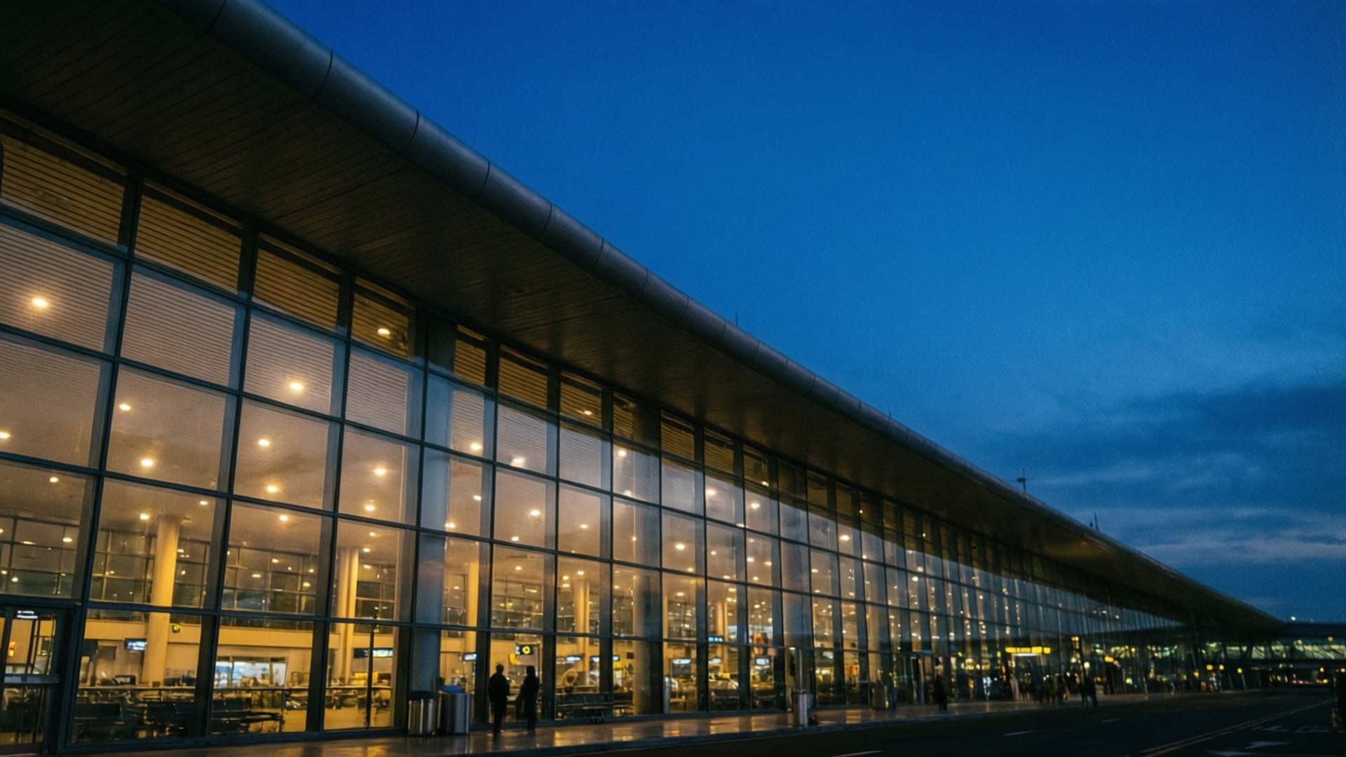 Blue Hour Terminal 壁紙 — 現代機場航廈在黃昏時溫暖地發光，玻璃外牆反射內部燈光，映襯著深藍色的天空。
