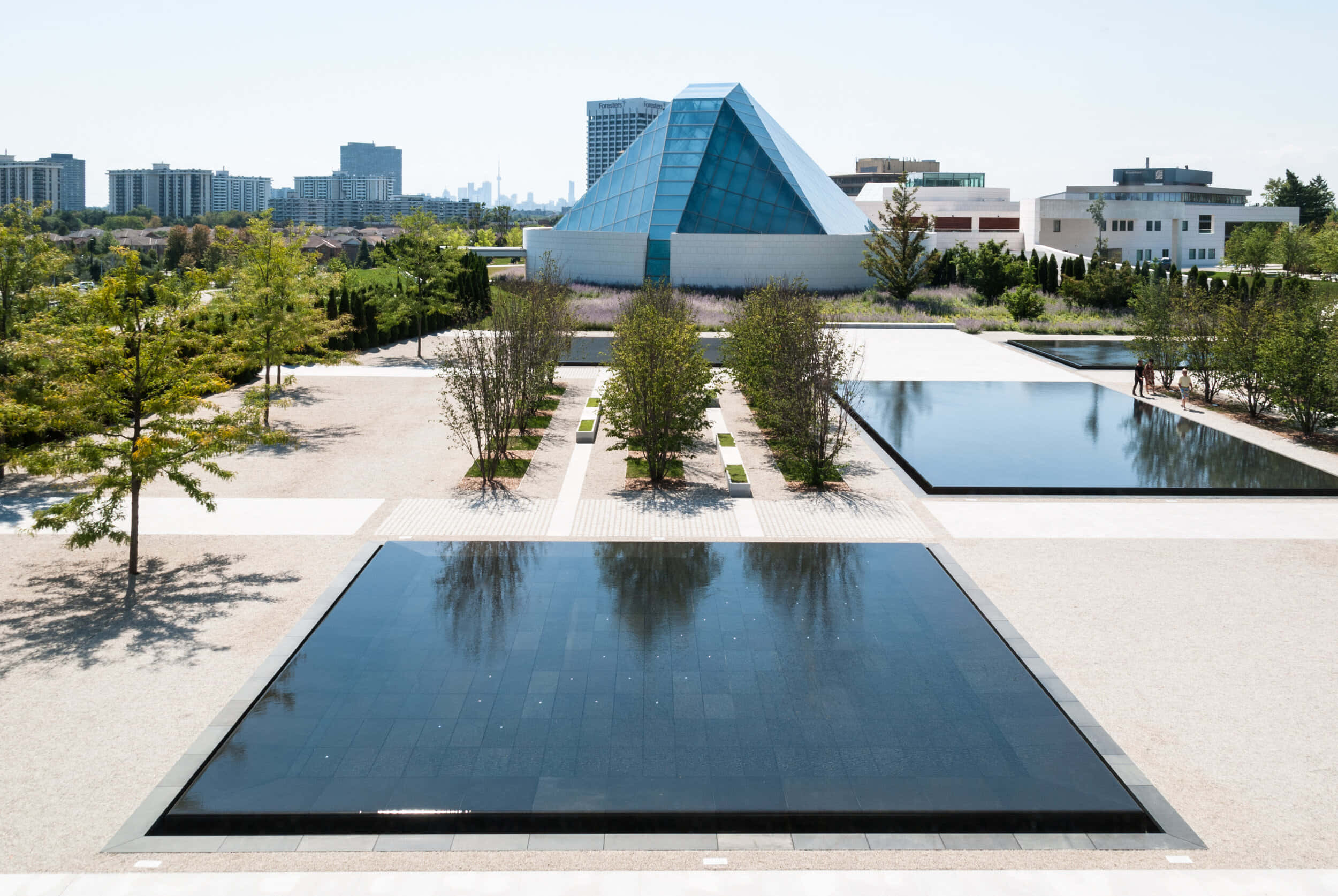 Aga Khan Museum 桌布 壁紙 — 一座現代玻璃金字塔建築在明亮的日光下，倒映在由樹木和城市天際線環繞的平靜長方形水池中。