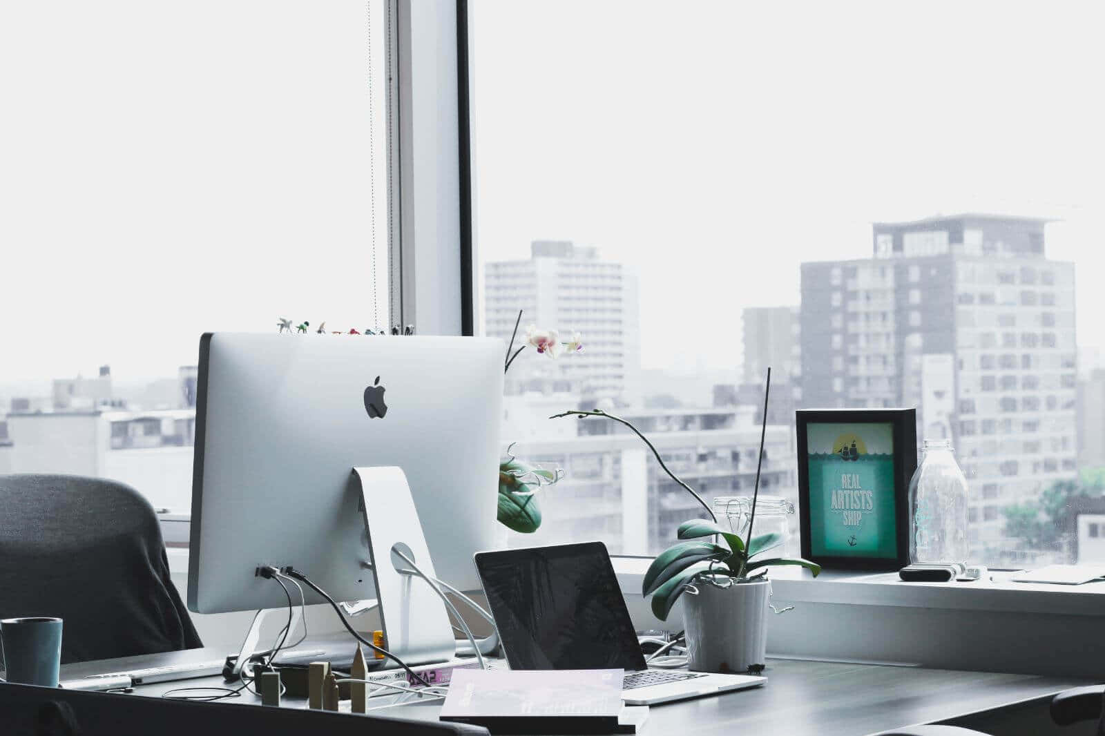  壁紙 — 現代辦公桌，配備 iMac 筆記型電腦和蘭花植物，透過大窗戶俯瞰明亮的城市景觀。