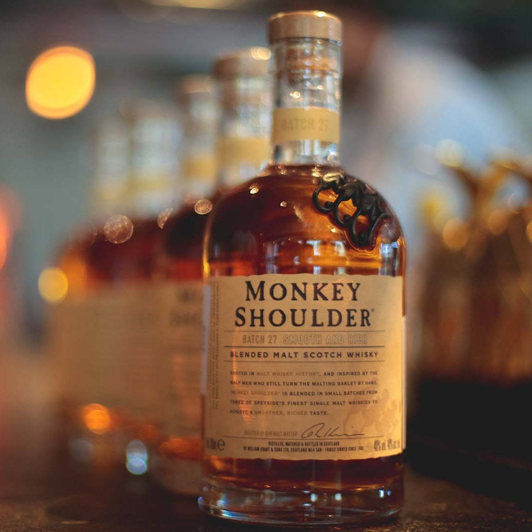 alcohol 桌布 壁紙 — Monkey Shoulder 威士忌酒瓶的特寫，在模糊的酒吧背景和金色散景燈光下溫暖地發光。