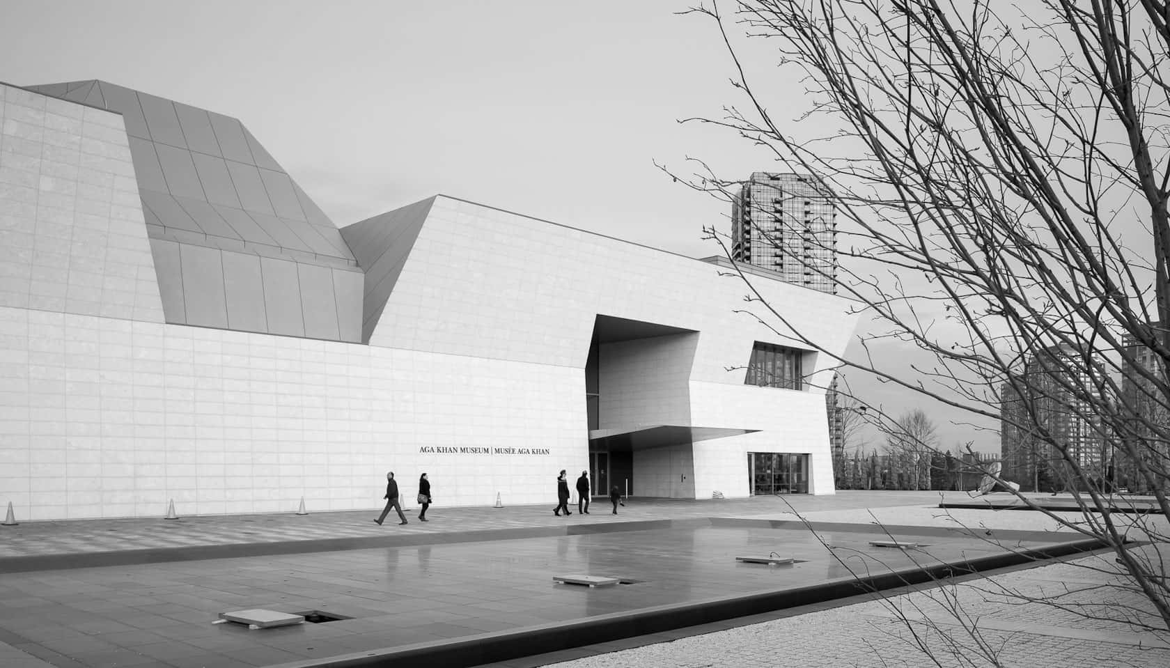 Aga Khan Museum 桌布 壁紙 — MCA Kiasan 博物館的單色建築拍攝，具有棱角分明的白色外牆和光禿禿的樹木，框住行人走在鋪砌的廣場上。