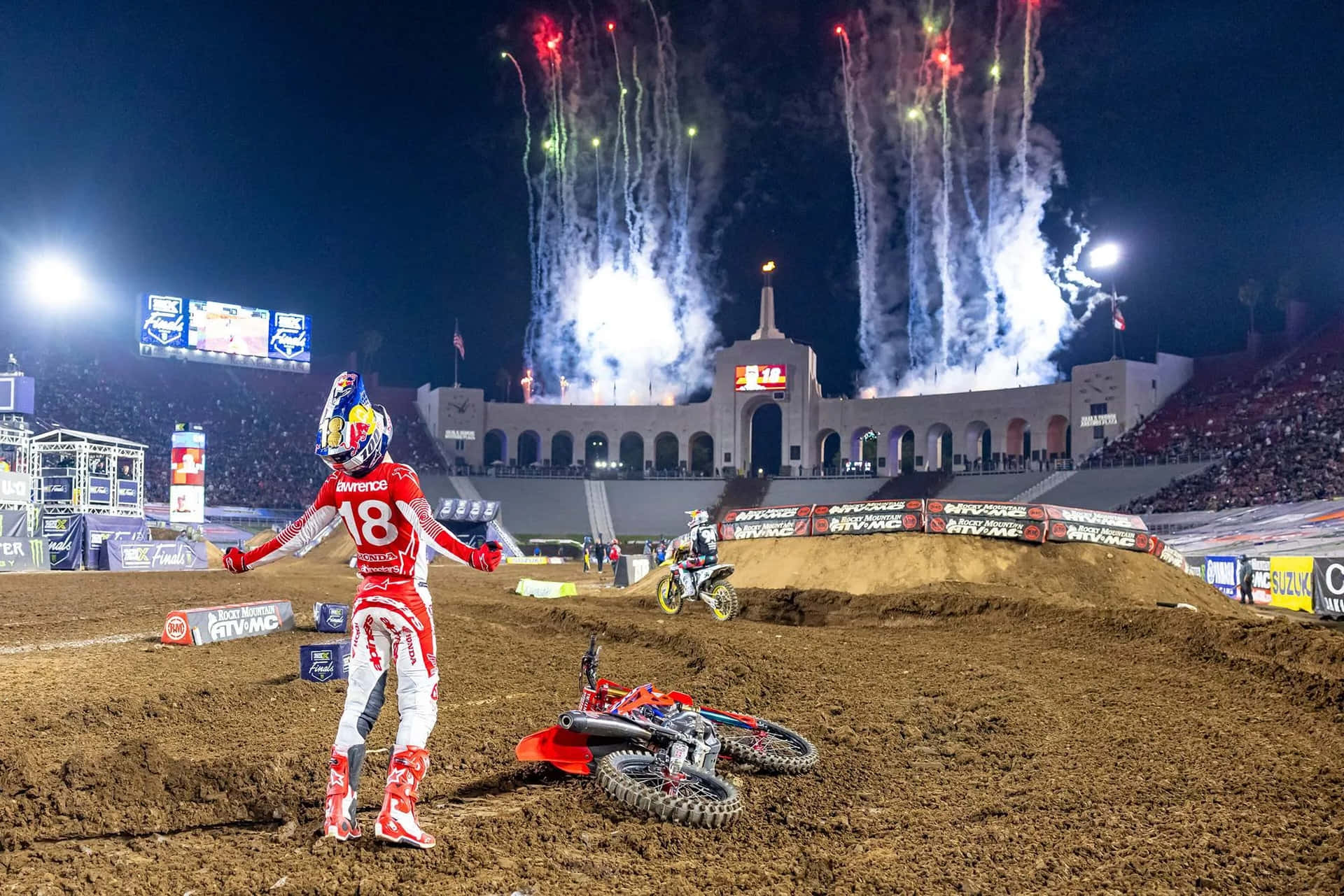  壁紙 — 一名身穿紅色裝備的越野摩托車手在夜晚慶祝勝利，煙火在擠滿觀眾的體育場上空綻放。