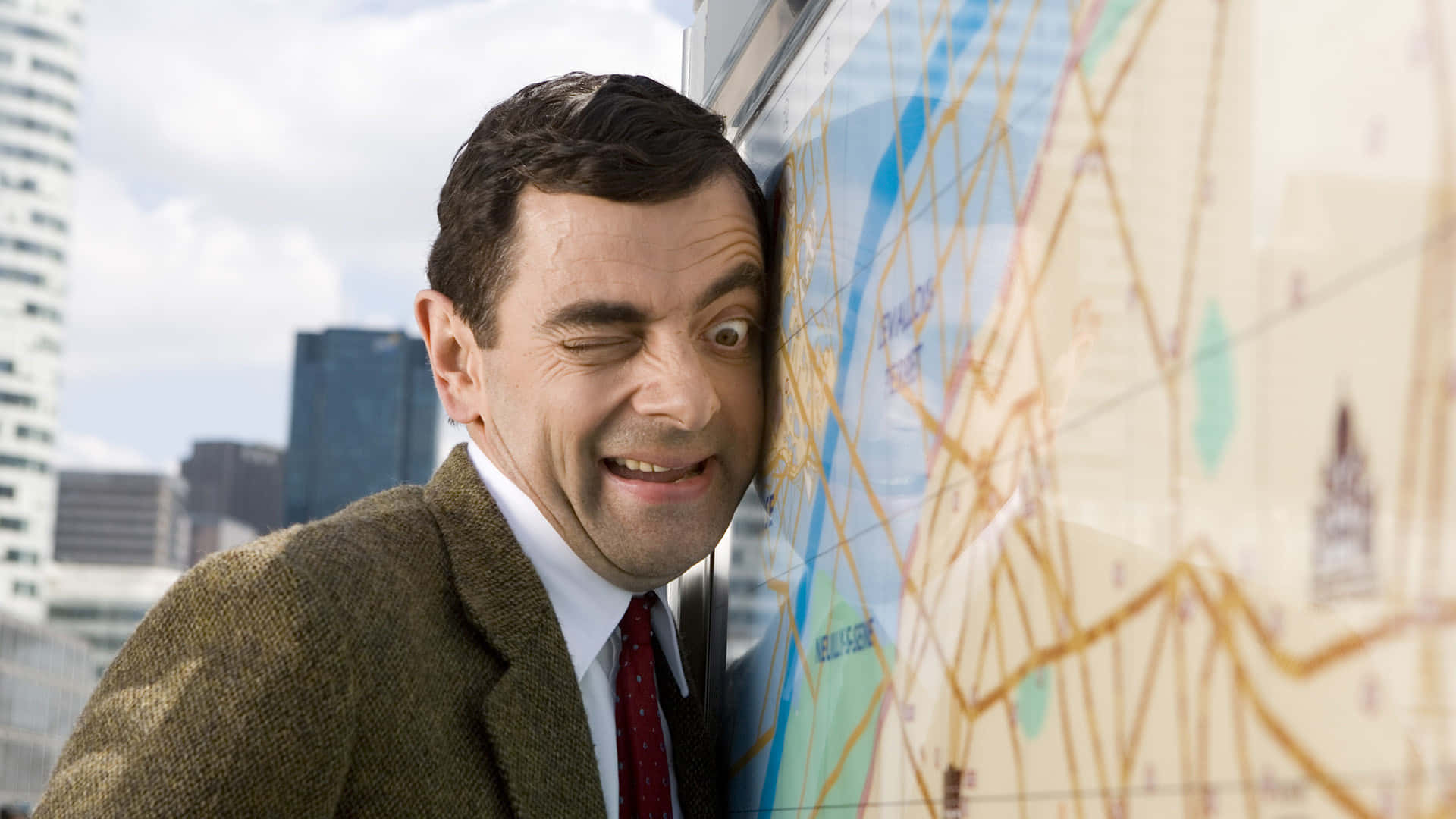 豆豆先生 桌布 壁紙 — Mr Bean 在一個大城市地圖後面偷看，背景有摩天大樓，露出頑皮的笑容。