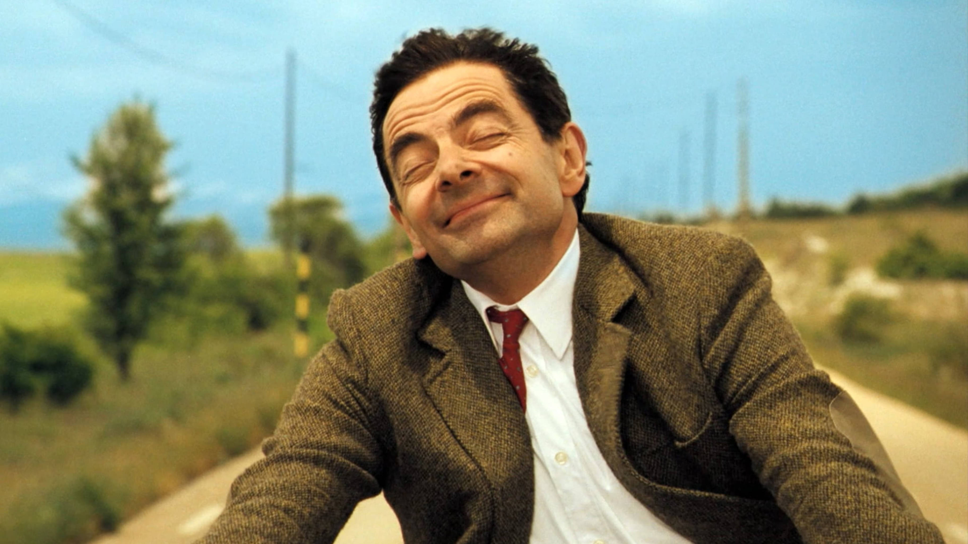 豆豆先生 桌布 壁紙 — Mr Bean 在晴朗的藍天下騎著腳踏車沿鄉村道路行駛，露出幸福的笑容。