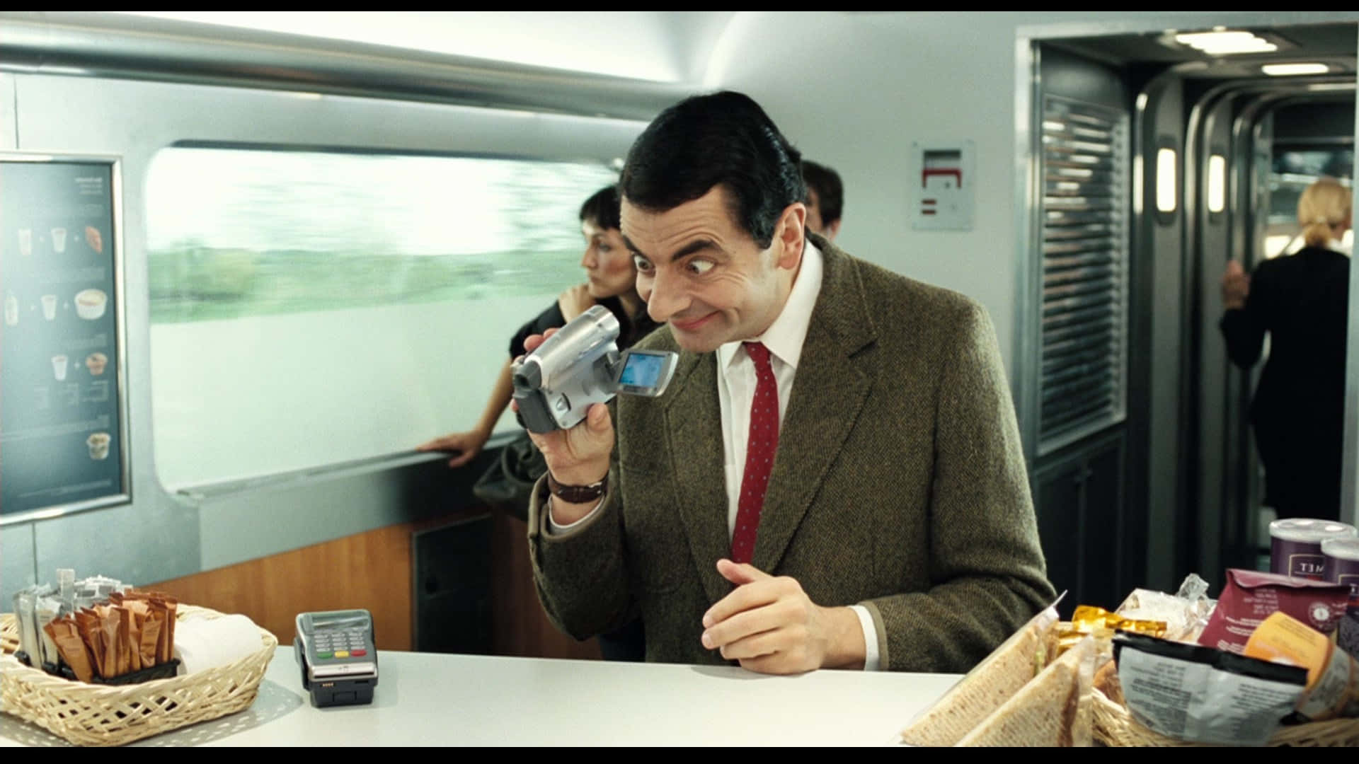 豆豆先生 桌布 壁紙 — Mr Bean 在火車車廂的小吃櫃檯前拿著一台攝影機，露出頑皮的笑容。