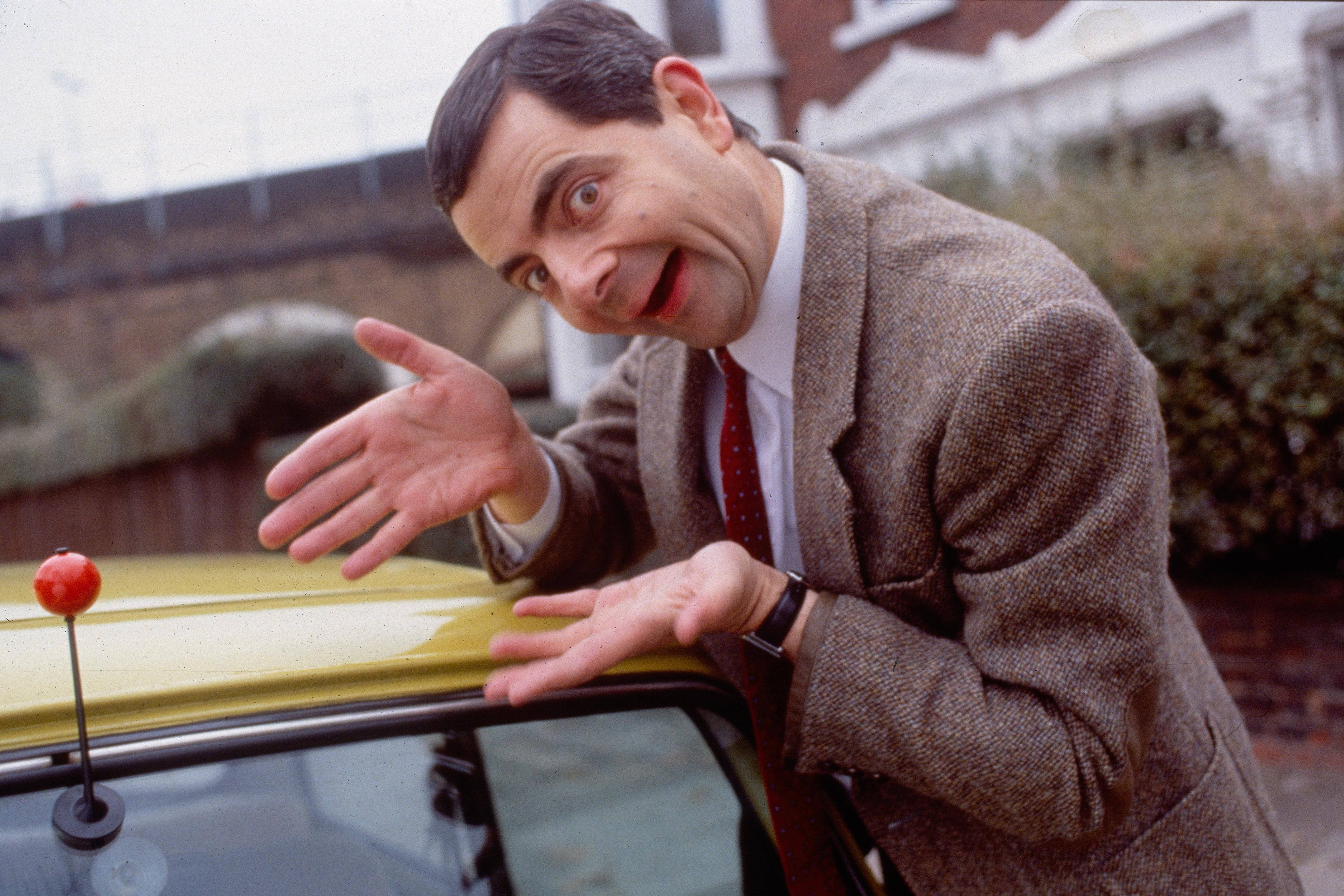 4K 桌布 壁紙 — Mr Bean 在郊區環境中俯身在一輛黃色車上，表情滑稽，雙手張開。