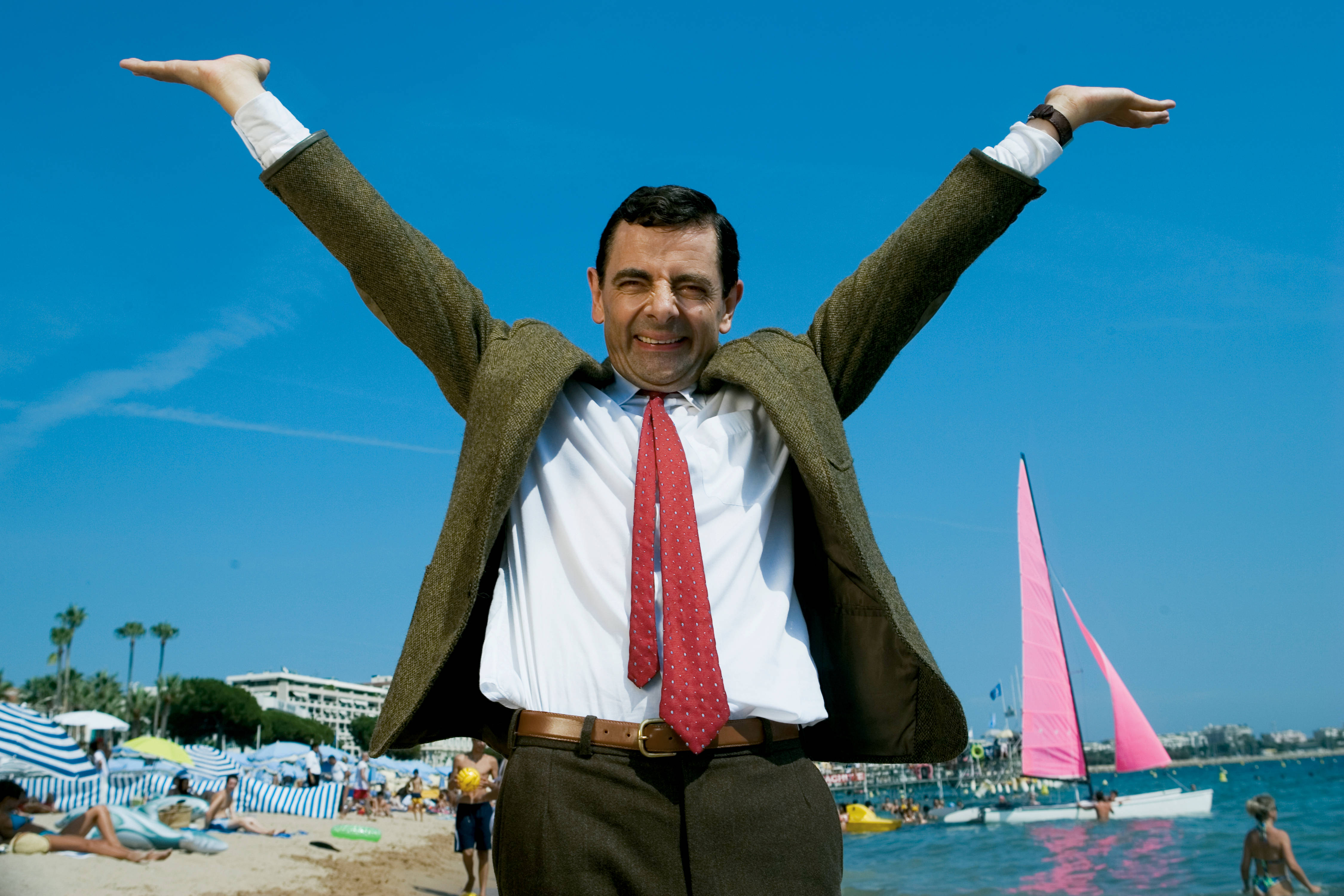 Mr Bean 在陽光明媚的海灘上，舉起雙臂，背景是粉紅色的帆船。 — 4K 壁紙