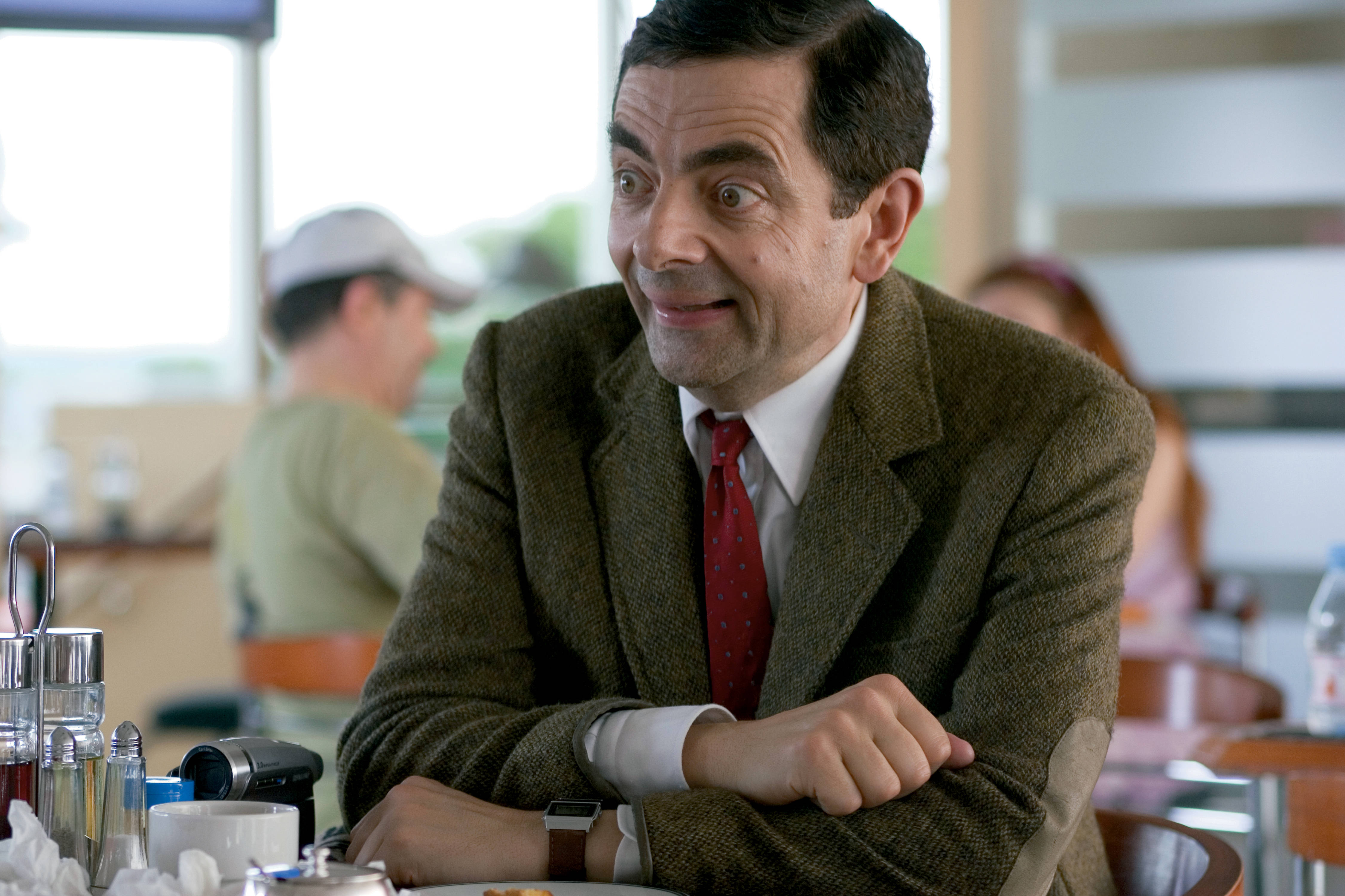 4K 桌布 壁紙 — Mr Bean 坐在咖啡廳桌子旁，睜大眼睛，露出頑皮的笑容，穿著棕色粗花呢外套和紅色領帶。