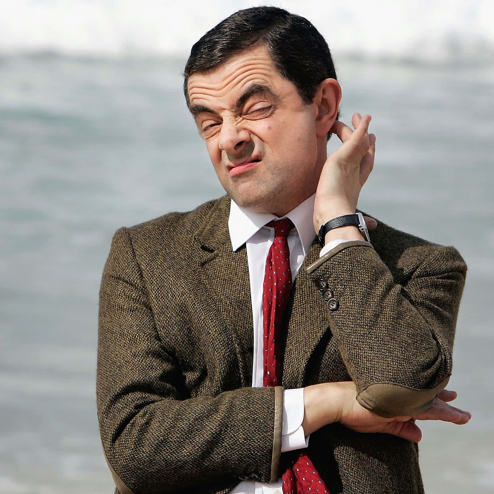 豆豆先生 桌布 壁紙 — Mr Bean 穿著粗花呢外套和紅色領帶，在水邊用滑稽誇張的表情抓耳朵。