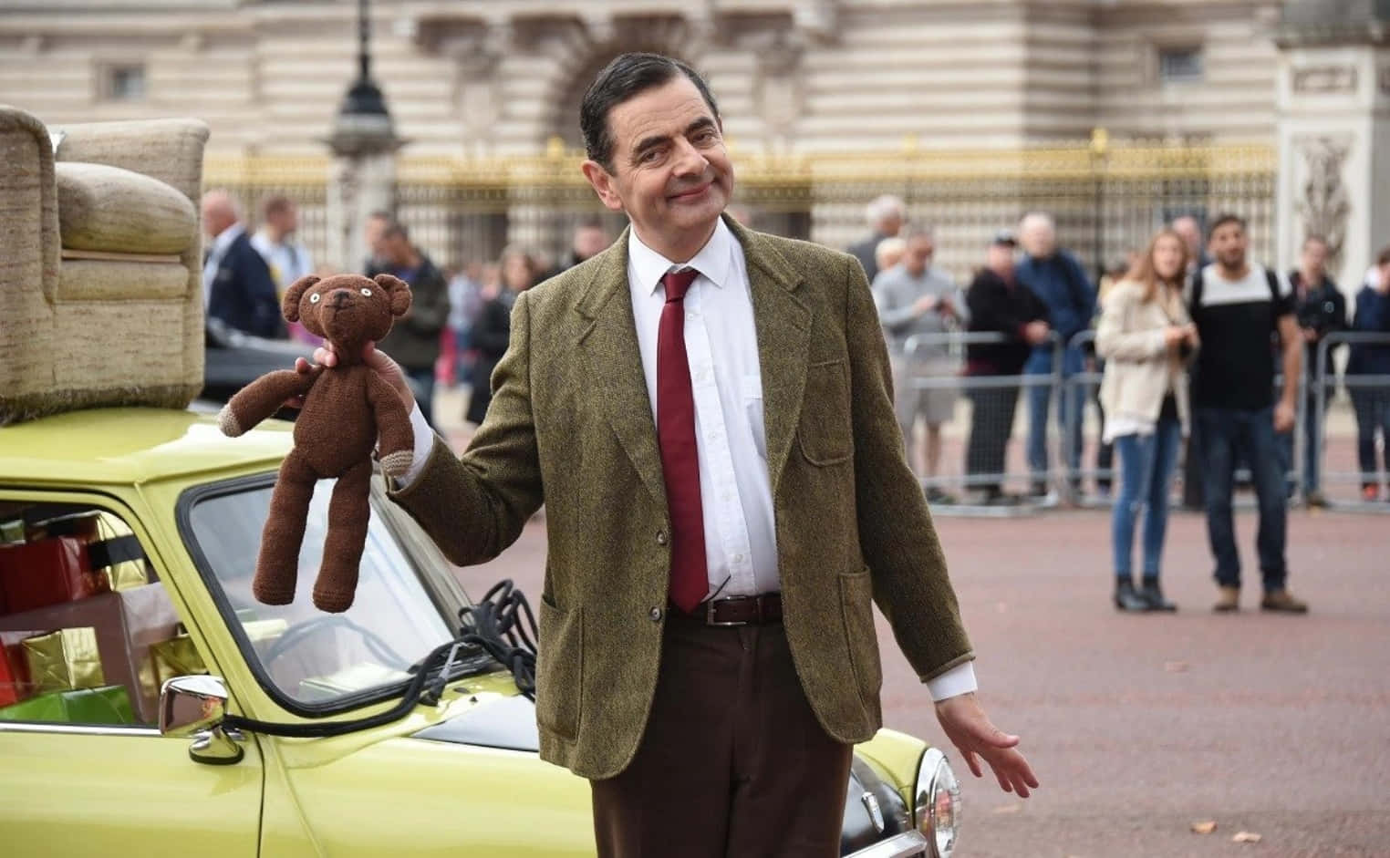 豆豆先生 桌布 壁紙 — Mr Bean 站在一輛黃色汽車旁邊，手持一隻泰迪熊，背後有一座宏偉的建築物。