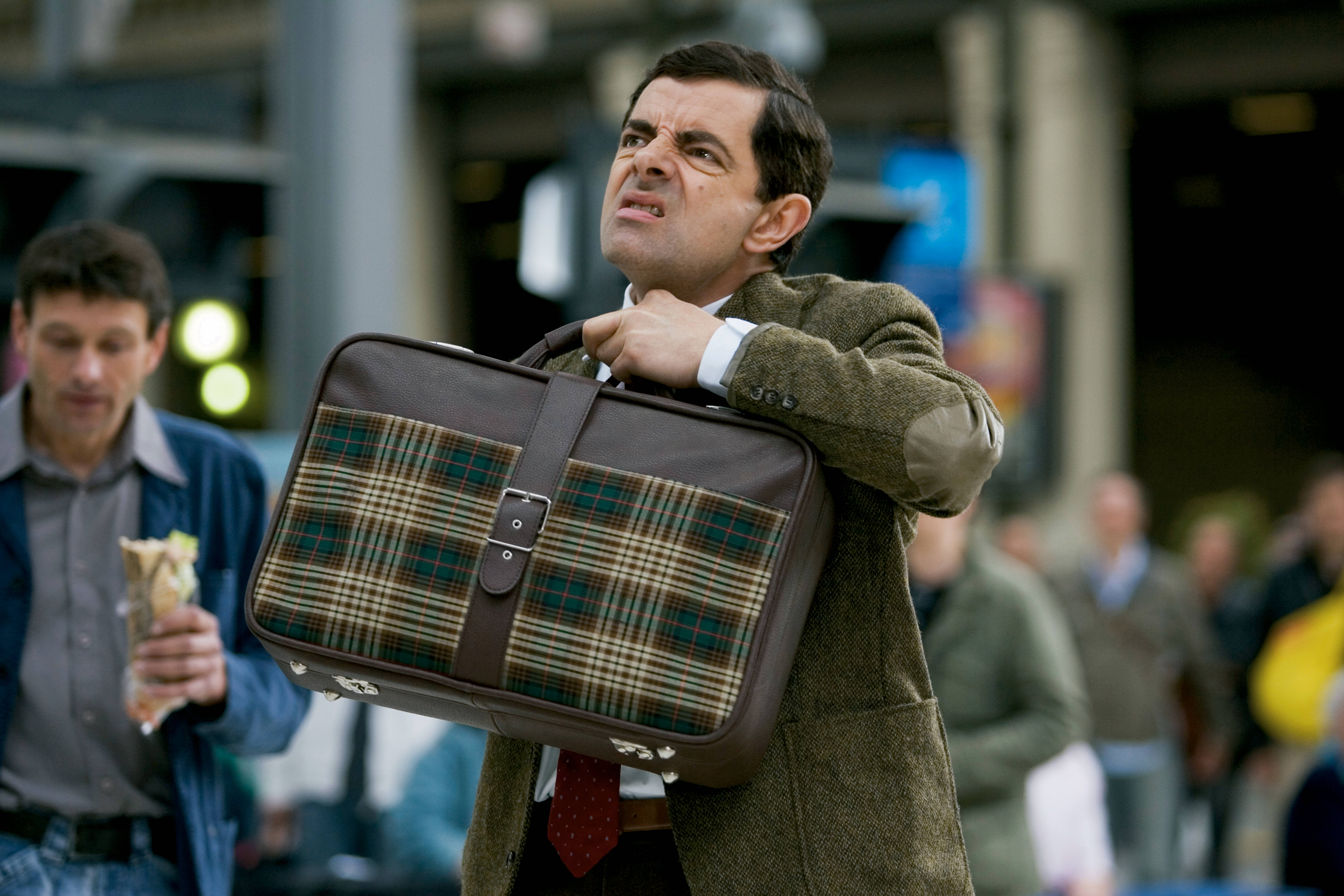 Mr Bean 在繁忙的車站努力拖著一個格子行李箱。 — 4K 壁紙