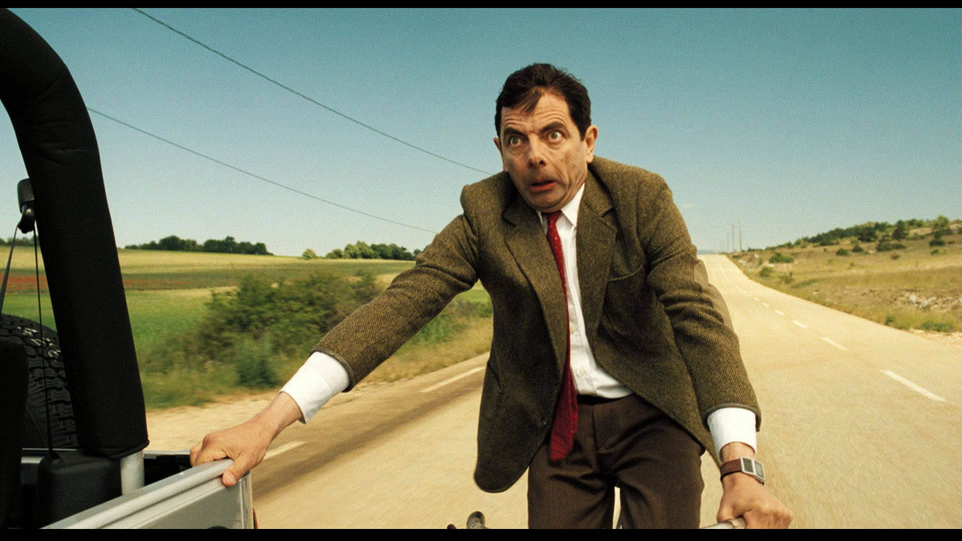 豆豆先生 桌布 壁紙 — Mr. Bean 身穿棕色西裝，在陽光明媚的鄉村道路上從一輛拋錨的汽車中探出身子，表情震驚。