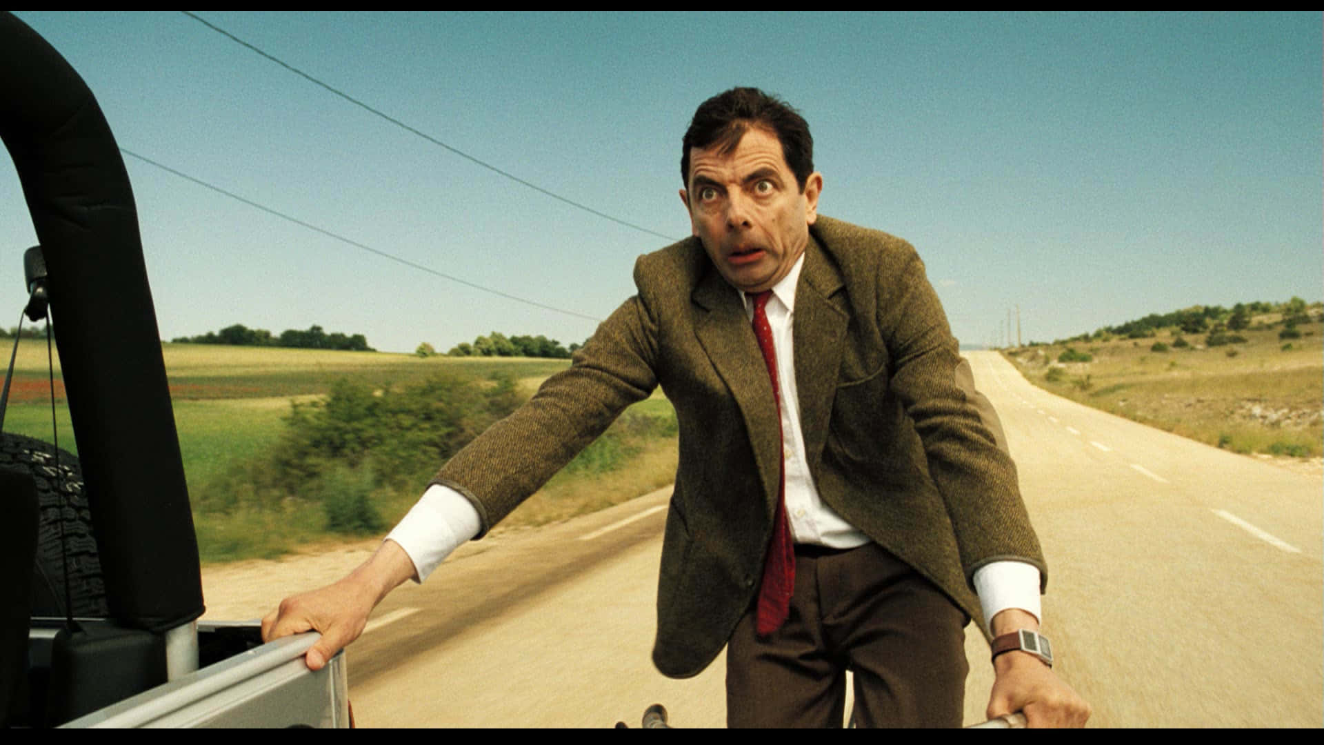 豆豆先生 桌布 壁紙 — Mr Bean 身穿棕色西裝，在陽光明媚的鄉村道路上從車內探出身子，表情震驚。