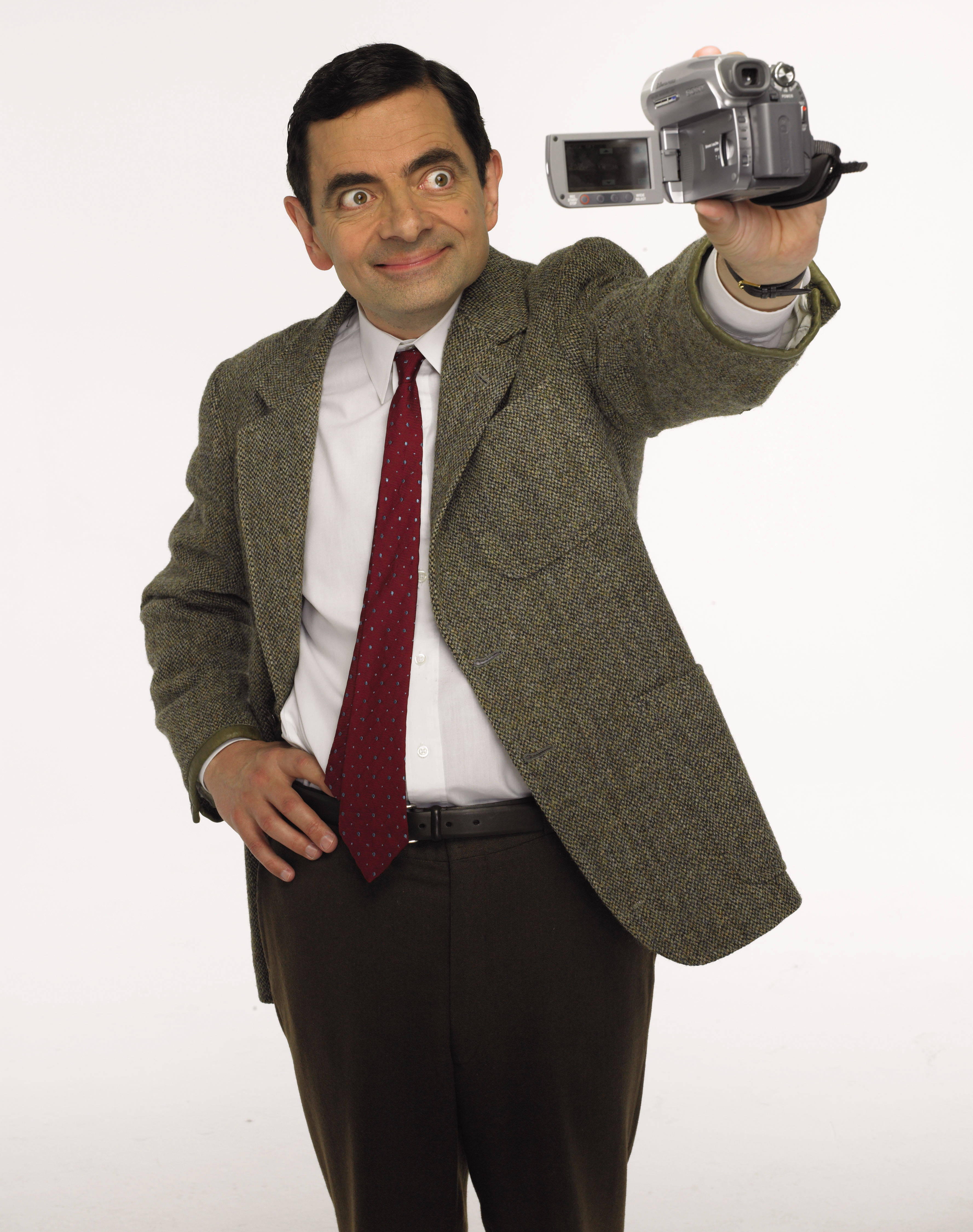 4K 桌布 壁紙 — Mr. Bean 身穿粗花呢外套和紅色領帶，手持攝錄機，睜大眼睛露出頑皮的笑容，背景為純白色。