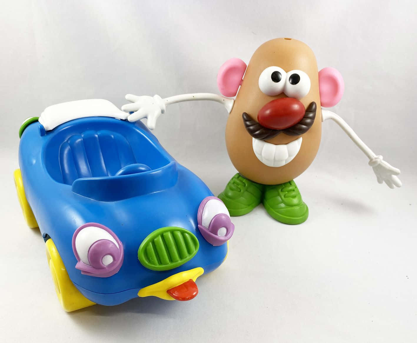 Mr Potato Head 桌布 壁紙 — Mr. Potato Head 有粉紅色耳朵和綠色鞋子，站在一輛藍色玩具車旁邊，車有黃色輪子和紫色眼睛。