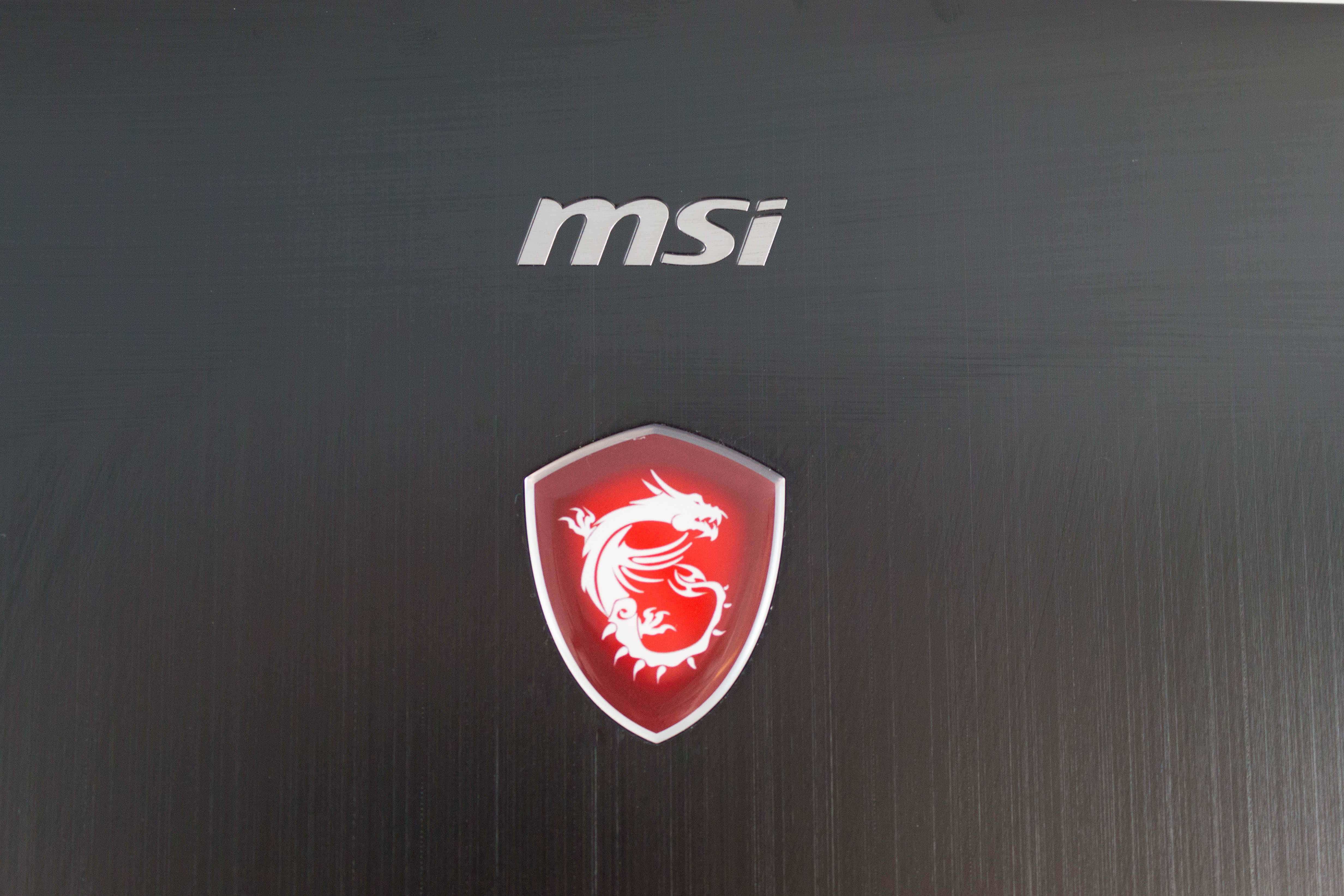 msi 2560x1080 桌布 壁紙 — MSI 筆記型電腦蓋的特寫，帶有發光的紅色龍標誌和銀色品牌標誌，位於拉絲黑色金屬上。
