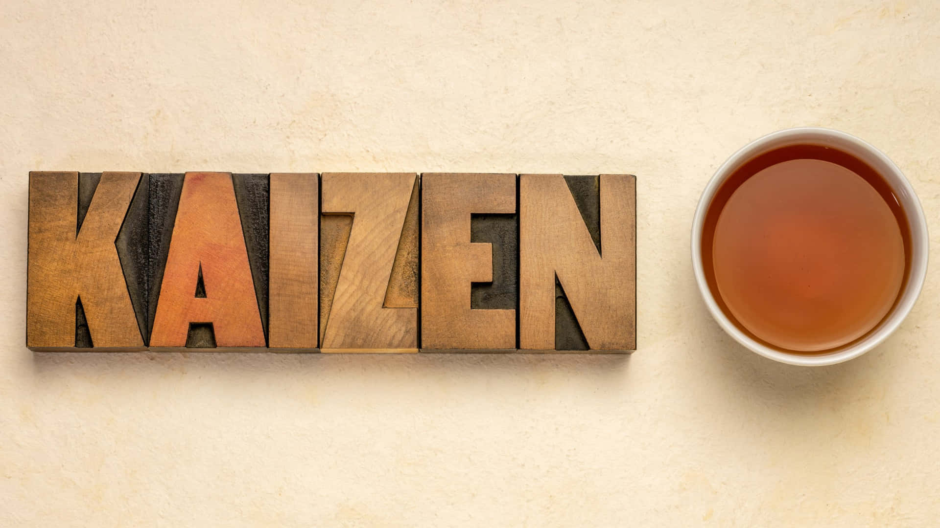  壁紙 — 木製活字塊拼出 kaizen，旁邊放著一杯琥珀色茶，置於有紋理的米色表面上。