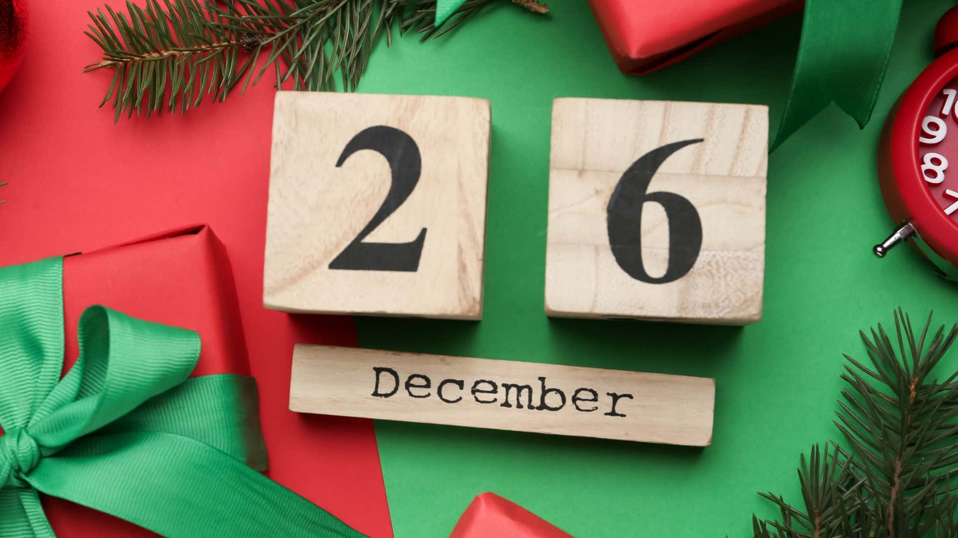  壁紙 — 木製日曆塊顯示12月26日，周圍環繞著紅色禮物、綠色蝴蝶結和松樹枝，背景為鮮豔的紅色和綠色。