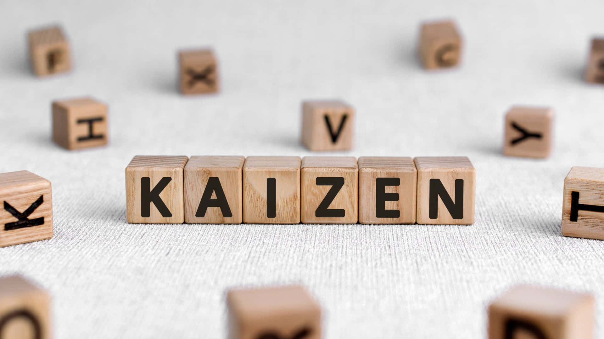  壁紙 — 木製字母塊在紋理灰色布料背景上清晰對焦拼出「KAIZEN」，周圍散落著字母塊。