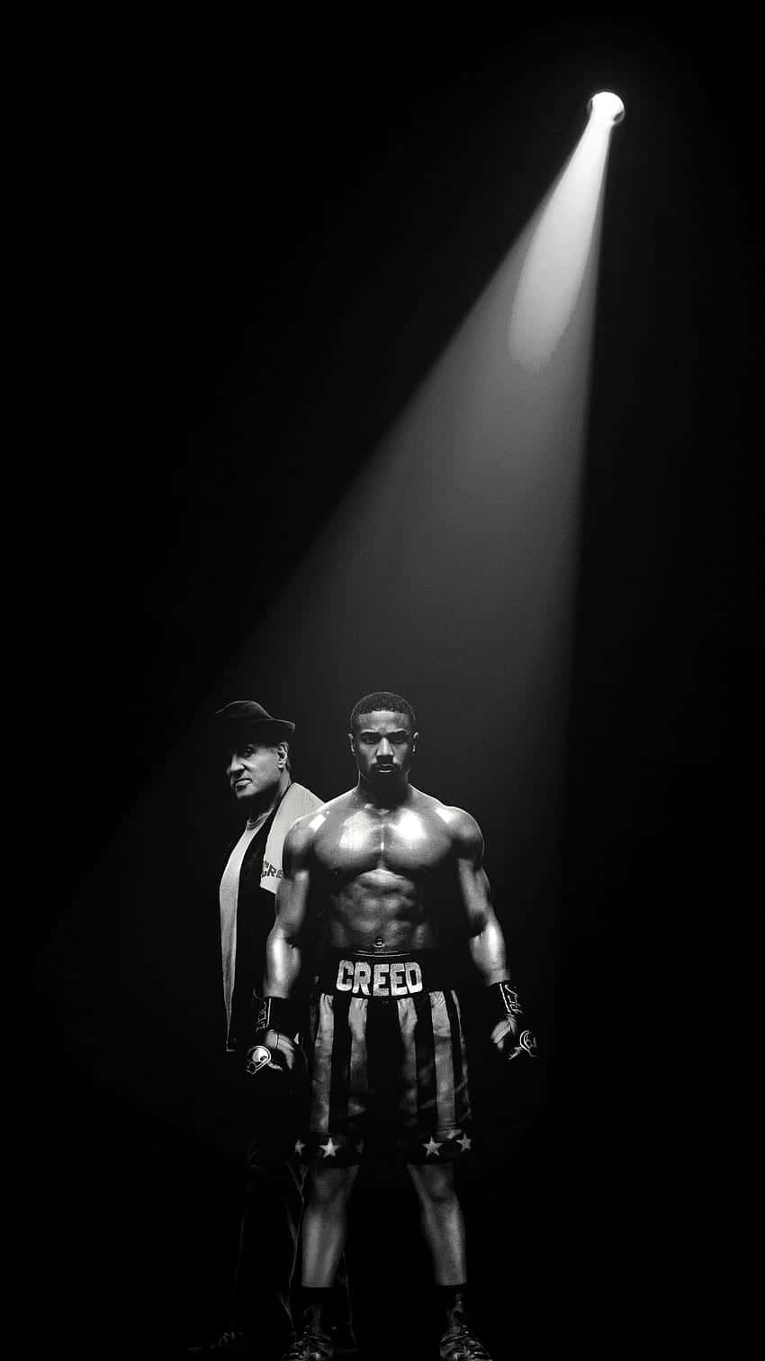  壁紙 — 一個肌肉發達的拳擊手穿著 'CREED' 短褲，站在單一聚光燈下，旁邊有一個戴帽子的男人，畫面是鮮明的黑白場景。