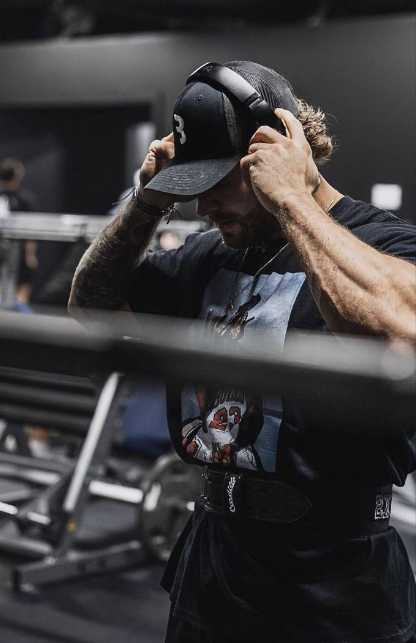 一個有刺青的男人在健身房裡，在舉重前調整耳機。 — Chris Bumstead 壁紙