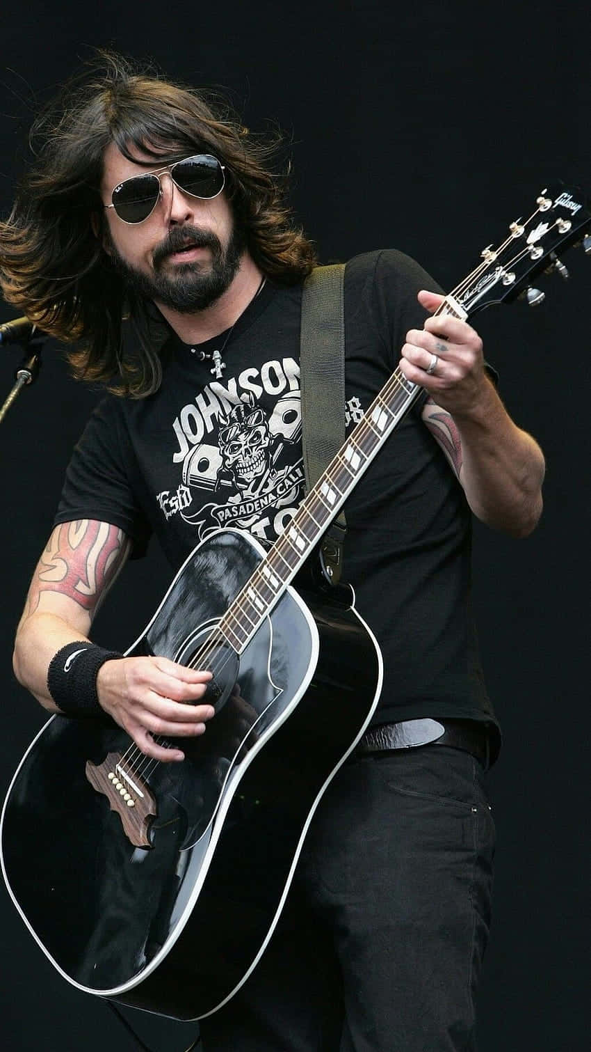 foo fighters 桌布 壁紙 — 一位長髮戴太陽眼鏡的音樂家在黑暗的舞台上熱情地彈奏黑色原聲吉他。
