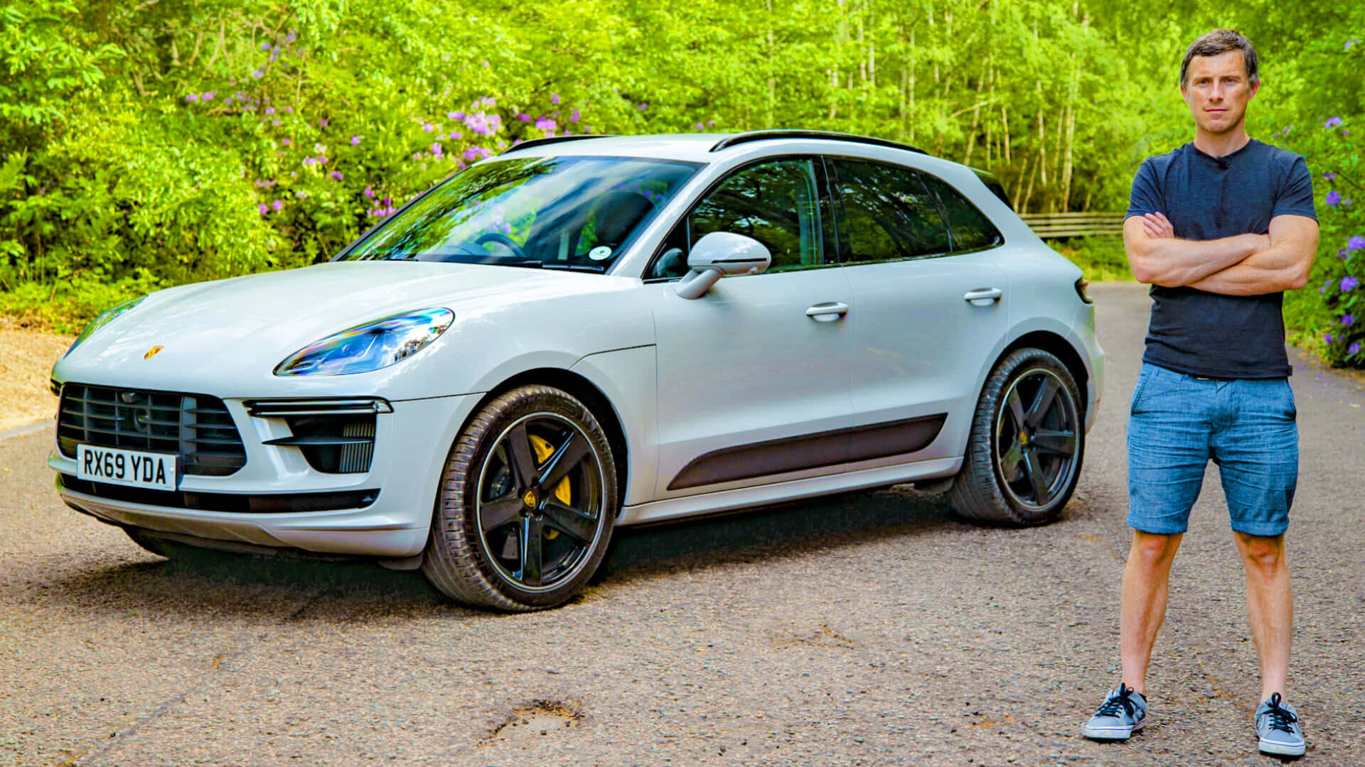 Porsche Macan 桌布 壁紙 — 一名男子自信地站在一輛停在碎石路上的時尚銀色保時捷 Macan SUV 旁邊，周圍環繞著茂密的綠植和紫色花朵。