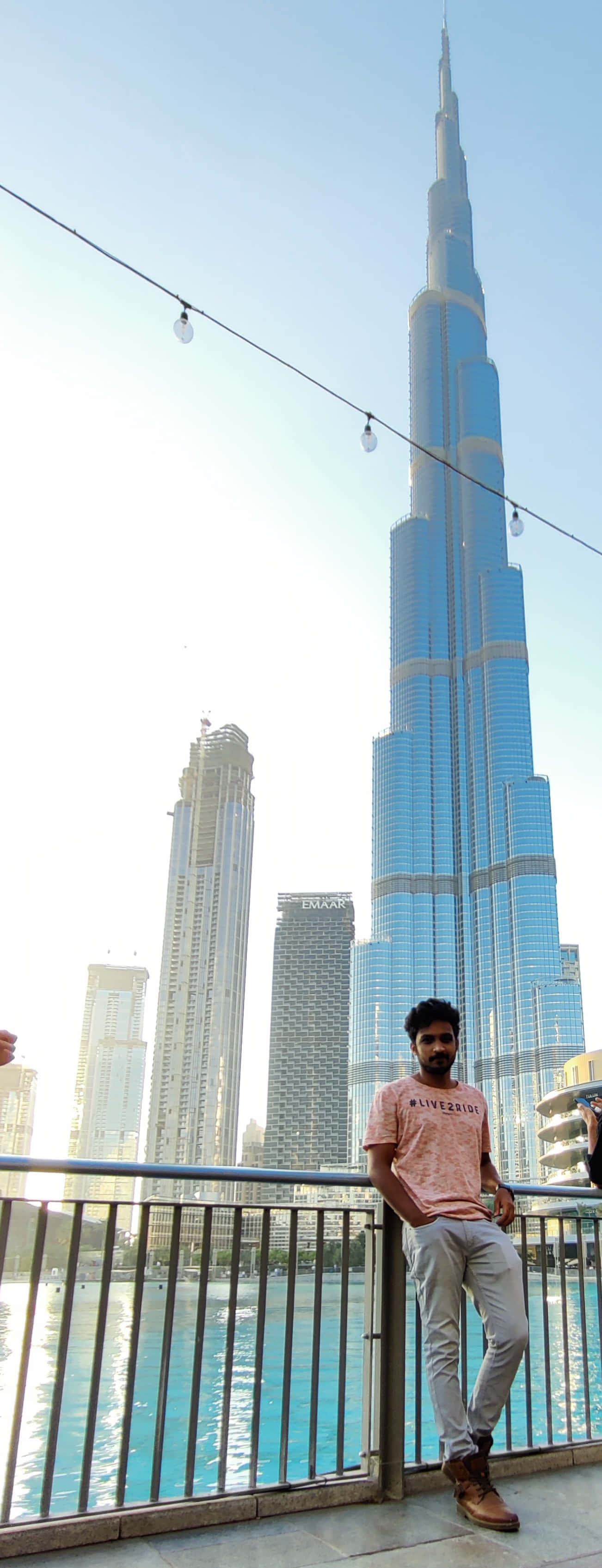 Burj Khalifa 桌布 壁紙 — 一名男子隨意靠在欄杆上，俯瞰水面，高聳的哈里發塔主宰了明亮的白天天際線。