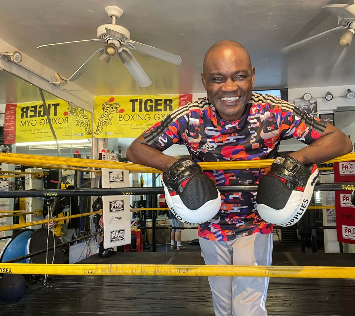  壁紙 — 一名微笑的男子穿著色彩繽紛的裝備，靠在健身房內掛有「Tiger Boxing Gym」橫幅的拳擊擂台繩上。