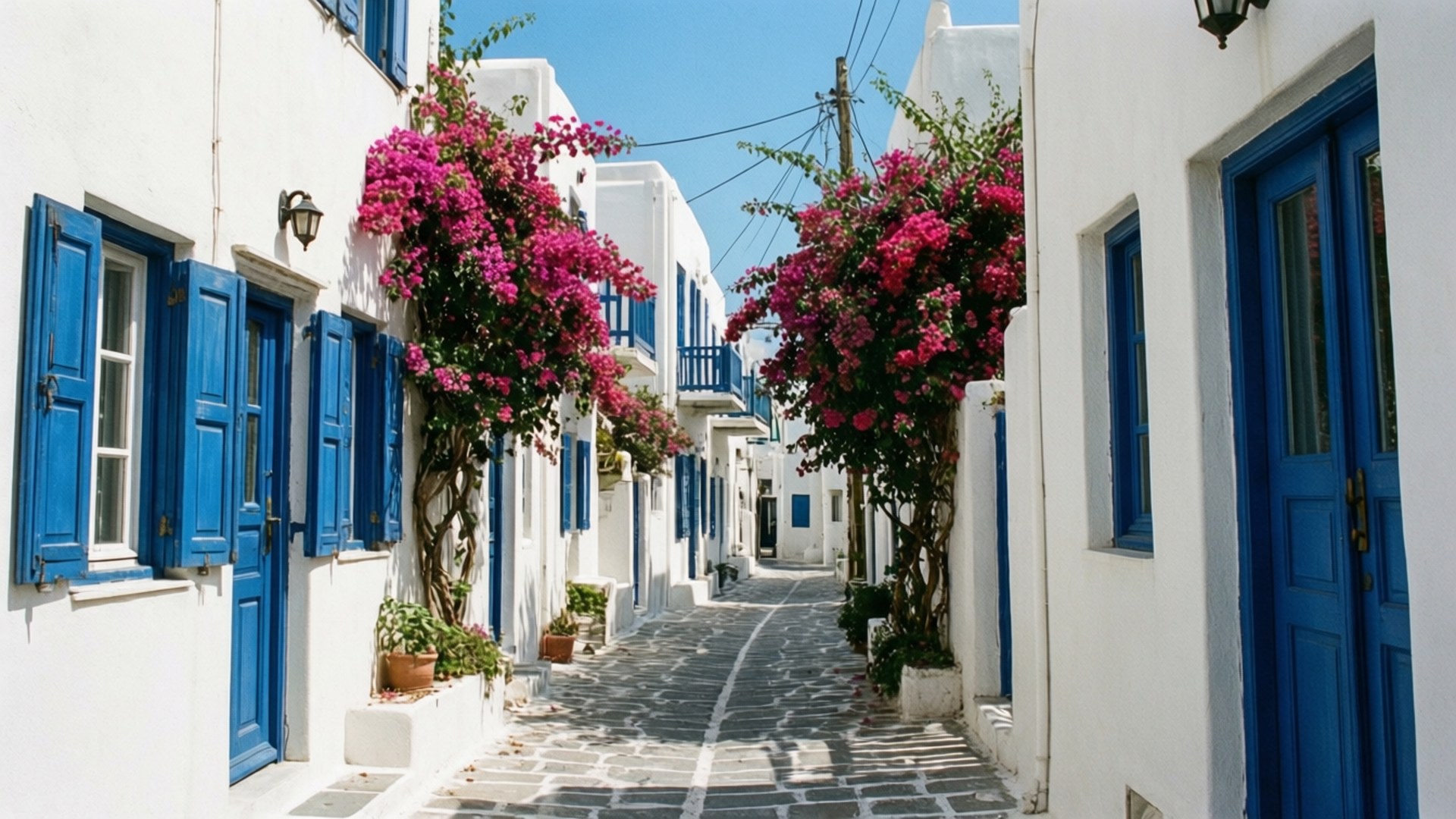 Mykonos Whitewashed Alley 壁紙 — 一條狹窄的鵝卵石小巷，兩旁是白色建築，鮮豔的粉紅色九重葛在藍色百葉窗上傾瀉而下，頭頂是清澈的藍天。