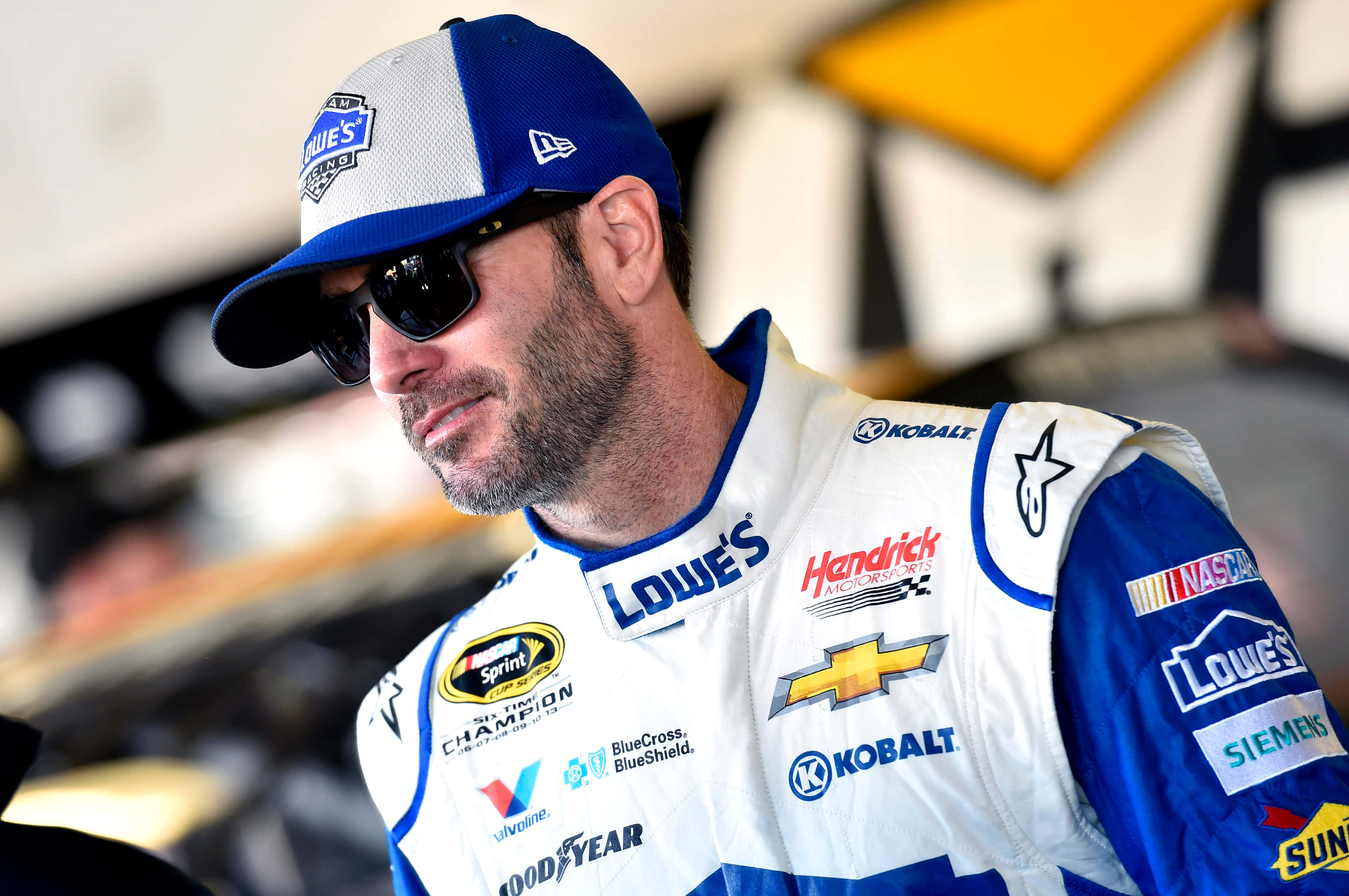 Jimmie Johnson 桌布 壁紙 — 一位身穿藍白賽車服、帶有贊助商標誌的NASCAR車手，戴著帽子和太陽眼鏡向下看。