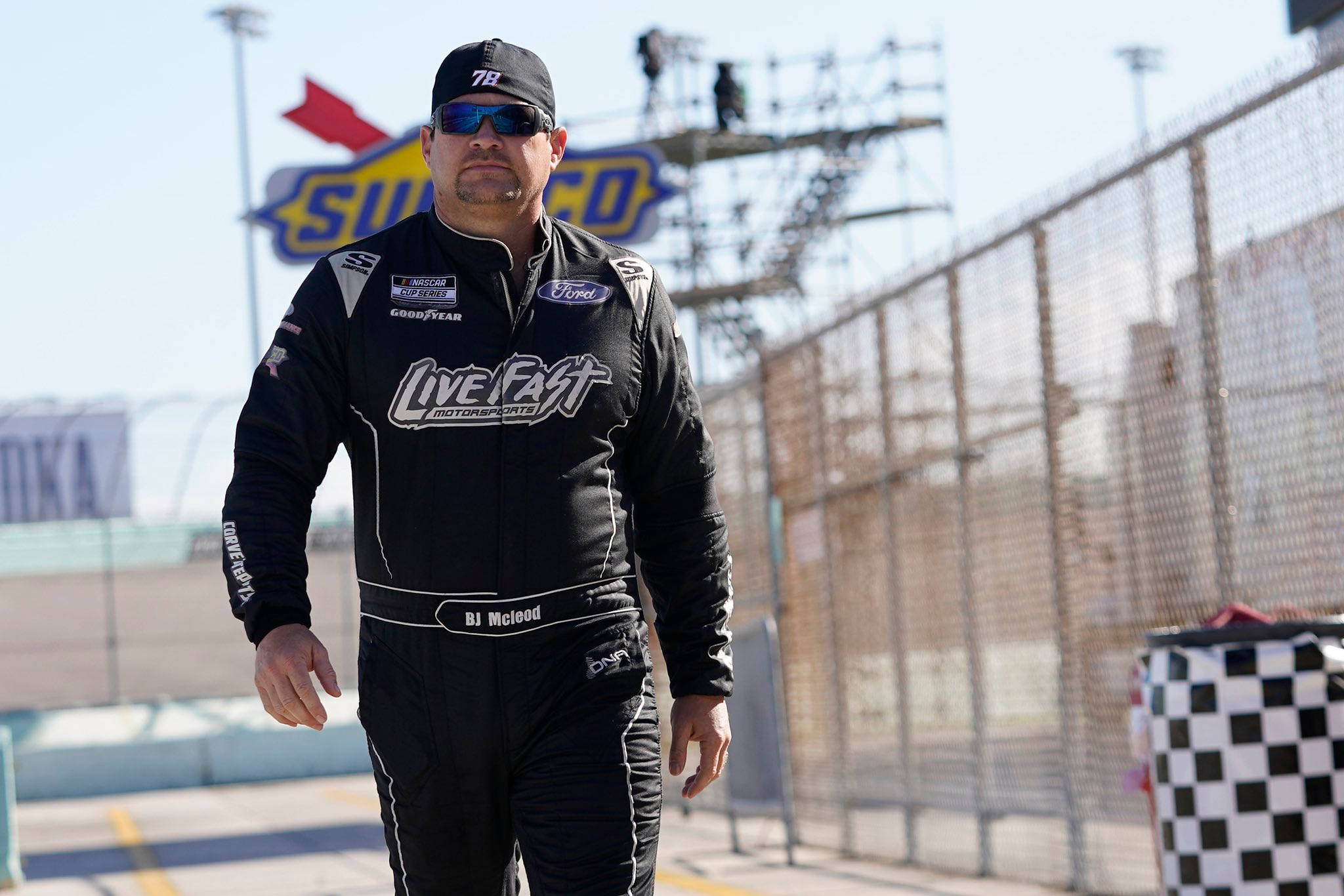 人物 桌布 壁紙 — NASCAR車手BJ McLeod身穿黑色賽車服，在陽光明媚的賽道上自信行走，背景有方格旗和'Sunoco'標誌。
