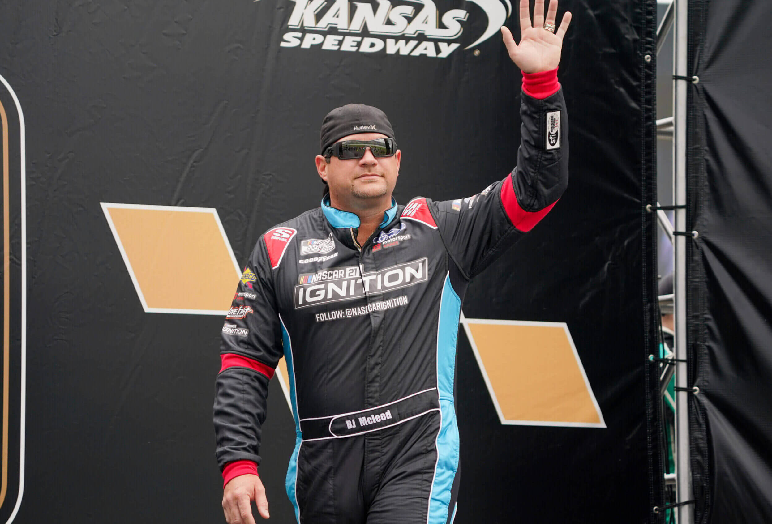 waving 桌布 壁紙 — NASCAR車手BJ McLeod在堪薩斯賽車場穿著黑色、藍色和紅色的Ignition賽車服向車迷揮手。